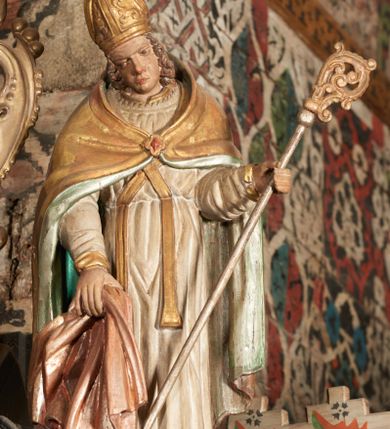 Zdjęcie nr 5: Rzeźba pełnoplastyczna, ścięta z tyłu, drążona. Święty przedstawiony en face, z pochyloną głową. Lewą ręką podtrzymuje pastorał, prawą kawałek pofałdowanej, różowej tkaniny. Twarz pociągła, o głęboko osadzonych oczach, duży nos. Włosy kręcone, opadają na ramiona. Święty ubrany w białą albę, skrzyżowaną na piersi stułę, przepasane w talii, oraz sztywną, złoconą kapę, podbitą na zielono, spiętą fibulą na piersi. Na głowie infuła biskupia. Polichromia naturalistczna, elementy stroju złocone i laserowane.