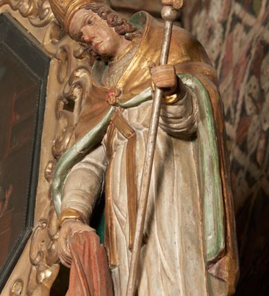 Zdjęcie nr 7: Rzeźba pełnoplastyczna, ścięta z tyłu, drążona. Święty przedstawiony en face, z pochyloną głową. Lewą ręką podtrzymuje pastorał, prawą kawałek pofałdowanej, różowej tkaniny. Twarz pociągła, o głęboko osadzonych oczach, duży nos. Włosy kręcone, opadają na ramiona. Święty ubrany w białą albę, skrzyżowaną na piersi stułę, przepasane w talii, oraz sztywną, złoconą kapę, podbitą na zielono, spiętą fibulą na piersi. Na głowie infuła biskupia. Polichromia naturalistczna, elementy stroju złocone i laserowane.