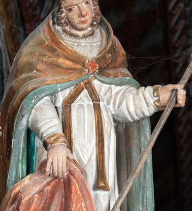 Zdjęcie nr 3: Rzeźba pełnoplastyczna, ścięta z tyłu, drążona. Święty przedstawiony en face, z pochyloną głową. Lewą ręką podtrzymuje pastorał, prawą kawałek pofałdowanej, różowej tkaniny. Twarz pociągła, o głęboko osadzonych oczach, duży nos. Włosy kręcone, opadają na ramiona. Święty ubrany w białą albę, skrzyżowaną na piersi stułę, przepasane w talii, oraz sztywną, złoconą kapę, podbitą na zielono, spiętą fibulą na piersi. Na głowie infuła biskupia. Polichromia naturalistczna, elementy stroju złocone i laserowane.