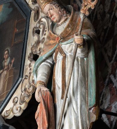 Zdjęcie nr 4: Rzeźba pełnoplastyczna, ścięta z tyłu, drążona. Święty przedstawiony en face, z pochyloną głową. Lewą ręką podtrzymuje pastorał, prawą kawałek pofałdowanej, różowej tkaniny. Twarz pociągła, o głęboko osadzonych oczach, duży nos. Włosy kręcone, opadają na ramiona. Święty ubrany w białą albę, skrzyżowaną na piersi stułę, przepasane w talii, oraz sztywną, złoconą kapę, podbitą na zielono, spiętą fibulą na piersi. Na głowie infuła biskupia. Polichromia naturalistczna, elementy stroju złocone i laserowane.