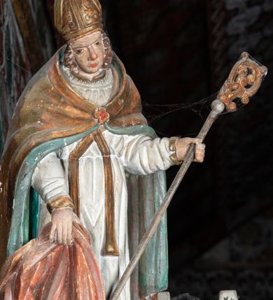 Zdjęcie nr 2: Rzeźba pełnoplastyczna, ścięta z tyłu, drążona. Święty przedstawiony en face, z pochyloną głową. Lewą ręką podtrzymuje pastorał, prawą kawałek pofałdowanej, różowej tkaniny. Twarz pociągła, o głęboko osadzonych oczach, duży nos. Włosy kręcone, opadają na ramiona. Święty ubrany w białą albę, skrzyżowaną na piersi stułę, przepasane w talii, oraz sztywną, złoconą kapę, podbitą na zielono, spiętą fibulą na piersi. Na głowie infuła biskupia. Polichromia naturalistczna, elementy stroju złocone i laserowane.