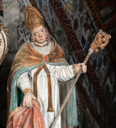 Zdjęcie nr 1: Rzeźba pełnoplastyczna, ścięta z tyłu, drążona. Święty przedstawiony en face, z pochyloną głową. Lewą ręką podtrzymuje pastorał, prawą kawałek pofałdowanej, różowej tkaniny. Twarz pociągła, o głęboko osadzonych oczach, duży nos. Włosy kręcone, opadają na ramiona. Święty ubrany w białą albę, skrzyżowaną na piersi stułę, przepasane w talii, oraz sztywną, złoconą kapę, podbitą na zielono, spiętą fibulą na piersi. Na głowie infuła biskupia. Polichromia naturalistczna, elementy stroju złocone i laserowane.