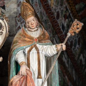 Zdjęcie nr 1: Rzeźba pełnoplastyczna, ścięta z tyłu, drążona. Święty przedstawiony en face, z pochyloną głową. Lewą ręką podtrzymuje pastorał, prawą kawałek pofałdowanej, różowej tkaniny. Twarz pociągła, o głęboko osadzonych oczach, duży nos. Włosy kręcone, opadają na ramiona. Święty ubrany w białą albę, skrzyżowaną na piersi stułę, przepasane w talii, oraz sztywną, złoconą kapę, podbitą na zielono, spiętą fibulą na piersi. Na głowie infuła biskupia. Polichromia naturalistczna, elementy stroju złocone i laserowane.