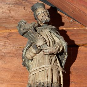 Zdjęcie nr 1: Rzeźba pełnoplastyczna, ustawiona na niewysokim cokole na rzucie prostokąta. Święty ukazany frontalnie, w kontrapoście, o esowato wygiętej sylwetce, z głową przechyloną na prawe ramię. Ręce ugięte w łokciach. Obiema dłońmi podtrzymuje przed sobą krucyfiks oparty na prawym ramieniu. Twarz pociągła, okolona zarostem; włosy krótkie. Ubrany w czarną sutannę, białą delikatnie drapowaną komżę, na ramionach mucet. Na głowie biret. Polichromia naturalistyczna.