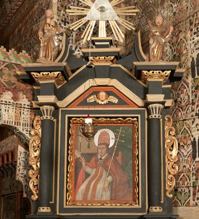 Zdjęcie nr 9: Obraz w kształcie stojącego prostokąta, w ozdobnej ramie. W centrum św. Grzegorz siedzący na tronie, ukazany do kolan, zwrócony en trois quarts w prawo; prawa ręka ugięta w łokciu z uniesioną otwartą dłonią, lewa wsparta na poręczy tronu z dłonią opuszczoną w dół podtrzymuje krzyż papieski. Twarz pociągła, o trójkątnej linii brody. Wyraźne rysy twarzy, oczy w kształcie migdałów, nos długi, prosty. Ubrany w obficie drapowaną w rurkowate fałdy rokietę o koronkowym zakończeniu rękawów. Na piersi czerwona stuła, na ramionach czerwona kapa o bogato zdobionej lamówce z przedstawieniem świętych, spięta na piersi dekoracyjną fibulą w formie kameryzowanej rozety. Na głowie tiara papieska. Wokół głowy nimb. W prawym górnym rogu obrazu podwieszona zielona kotara. W tle po lewej stronie stół z pasyjką oraz otwartą księgą. Na stole napis: S. ALEKSANDER. Kolorystyka ciemna, przytłumiona z przewagą brązów i czerwieni. Linearyzm, formy wydobyte konturem.