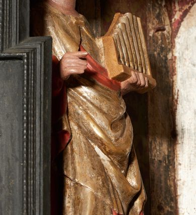 Zdjęcie nr 19: Retabulum ołtarzowe z dużą rzeźbą Matki Boskiej z Dzieciątkiem oraz mniejszymi o połowę czterema rzeźbami Świętych Dziewic umieszczonymi w prostopadłościennej szafie na rzucie trapezu (z ukośnie ustawionymi ściankami bocznymi). Rzeźby pełnoplastyczne, ścięte z tyłu, drążone, ustawione na cokolikach. Pośrodku figura Matki Boskiej w stylu gotyku międzynarodowego; zwrócona en face, w delikatnym kontrapoście, o esowato wygiętej sylwetce, podtrzymuje na obu rękach wpółleżące Dzieciatko. Twarz owalna z podwójnym podbródkiem, rysy twarzy płytko rzeźbione, delikatne, długi, prosty nos. Włosy upięte z tyłu. Maria ubrana w srebrzoną suknię z tkaniną drapowaną po środku w trzy fałdy misowe oraz złocony płaszcz podbity na niebieską podszewką, spływający od tyłu głowy na ramiona i boki, w układzie kaskadowym. Na głowie korona otwarta. Dzieciatko wpółleżące, zwrócone en trois quarts w prawo, prawą ręka ujmuje Matkę za szyję, w lewej trzyma złotą kulę. Twarz okrągła, duże uszy, włosy krótkie, kręcone. W partii bioder przepasany ściśle przylegającą, złotą tkaniną, delikatnie drapowaną. Na lewej ściance szafy u góry figurka św. Cecylii, zwróconej en face, o esowato wygiętej sylwetce, w lewej ręce, opartej na biodrze trzyma piszczałki, prawa wyciągnięta do przodu. Twarz okrągła o pełnych policzkach i płytko rzeźbionych rysach twarzy, włosy długie, falujące fakturowane, spływające przez ramiona na plecy. Ubrana w srebrzoną suknię oraz złocony płaszcz z czerwoną podszewką, spięty na piersi fibulą, z obfitą draperią przewieszoną przez ramiona, opadająca na froncie fałdami misowymi oraz spływającymi kaskadami od ramion. Na głowie blok drewna z namalowaną koroną otwartą.Na lewej ściance u dołu figura św. Katarzyny Aleksandryjskiej, zwróconej en face, w prawej ręce podtrzymuje koło, lewą wyciąga do przodu. Twarz okrągła, policzki pełne, o wysokim czole, nos duży, oczy duże delikatnie zarysowane. Włosy falowane, fakturowane, spływają na plecy. Święta ubrana w srebrzona suknię przepasaną w talii oraz złocony płaszcz spięty broszą na piersi, z obfitą draperią przewieszoną przez ramiona, opadająca na froncie misowymi fałdami i spływającymi kaskadami od ramion. Na głowie blok drewna z namalowaną koroną otwartą. Na prawej ściance u góry figura św. Doroty, zwrócona en face w delikatnym kontrapoście, prawa ręką podtrzymuje koszyk, lewą wskazuje na niego. Twarz owalna, pełna, delikatne rysy, wąski zadarty nos. Włosy falowane, fakturowane, opadają na tył głowy. Ubrana w złotą suknię, delikatnie drapowaną u dołu, oraz srebrzony płaszcz podbity czerwoną podszewką, spięty na piersi i układający się z przodu w fałdy misowe. Na głowie blok drewna z namalowaną koroną otwartą. Na prawej ściance u dołu figura św. Barbary, zwróconej en face, o esowato przegiętej sylwetce, obiema rękami przytrzymuje opartą na lewym ramieniu wieżę. Twarz owalna, pełna, delikatne rysy, wąski zadarty nos. Włosy falowane, fakturowane, opadają na tył głowy. Ubrana w suknię i srebrzony płaszcz z niebieską podszewką, spięty na piersi fibulą, przerzucony przez ramiona z obfitą partią materiału układającą się z przodu w fałdy misowe. Na głowie blok drewna z namalowaną koroną otwartą. Karnacje w tonacji naturalnej, szaty srebrzone i złocone.