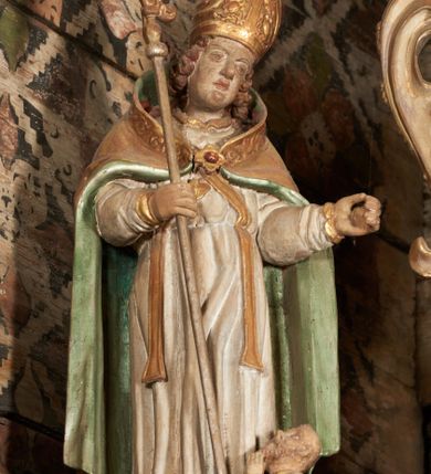 Zdjęcie nr 34: Retabulum ołtarzowe z dużą rzeźbą Matki Boskiej z Dzieciątkiem oraz mniejszymi o połowę czterema rzeźbami Świętych Dziewic umieszczonymi w prostopadłościennej szafie na rzucie trapezu (z ukośnie ustawionymi ściankami bocznymi). Rzeźby pełnoplastyczne, ścięte z tyłu, drążone, ustawione na cokolikach. Pośrodku figura Matki Boskiej w stylu gotyku międzynarodowego; zwrócona en face, w delikatnym kontrapoście, o esowato wygiętej sylwetce, podtrzymuje na obu rękach wpółleżące Dzieciatko. Twarz owalna z podwójnym podbródkiem, rysy twarzy płytko rzeźbione, delikatne, długi, prosty nos. Włosy upięte z tyłu. Maria ubrana w srebrzoną suknię z tkaniną drapowaną po środku w trzy fałdy misowe oraz złocony płaszcz podbity na niebieską podszewką, spływający od tyłu głowy na ramiona i boki, w układzie kaskadowym. Na głowie korona otwarta. Dzieciatko wpółleżące, zwrócone en trois quarts w prawo, prawą ręka ujmuje Matkę za szyję, w lewej trzyma złotą kulę. Twarz okrągła, duże uszy, włosy krótkie, kręcone. W partii bioder przepasany ściśle przylegającą, złotą tkaniną, delikatnie drapowaną. Na lewej ściance szafy u góry figurka św. Cecylii, zwróconej en face, o esowato wygiętej sylwetce, w lewej ręce, opartej na biodrze trzyma piszczałki, prawa wyciągnięta do przodu. Twarz okrągła o pełnych policzkach i płytko rzeźbionych rysach twarzy, włosy długie, falujące fakturowane, spływające przez ramiona na plecy. Ubrana w srebrzoną suknię oraz złocony płaszcz z czerwoną podszewką, spięty na piersi fibulą, z obfitą draperią przewieszoną przez ramiona, opadająca na froncie fałdami misowymi oraz spływającymi kaskadami od ramion. Na głowie blok drewna z namalowaną koroną otwartą.Na lewej ściance u dołu figura św. Katarzyny Aleksandryjskiej, zwróconej en face, w prawej ręce podtrzymuje koło, lewą wyciąga do przodu. Twarz okrągła, policzki pełne, o wysokim czole, nos duży, oczy duże delikatnie zarysowane. Włosy falowane, fakturowane, spływają na plecy. Święta ubrana w srebrzona suknię przepasaną w talii oraz złocony płaszcz spięty broszą na piersi, z obfitą draperią przewieszoną przez ramiona, opadająca na froncie misowymi fałdami i spływającymi kaskadami od ramion. Na głowie blok drewna z namalowaną koroną otwartą. Na prawej ściance u góry figura św. Doroty, zwrócona en face w delikatnym kontrapoście, prawa ręką podtrzymuje koszyk, lewą wskazuje na niego. Twarz owalna, pełna, delikatne rysy, wąski zadarty nos. Włosy falowane, fakturowane, opadają na tył głowy. Ubrana w złotą suknię, delikatnie drapowaną u dołu, oraz srebrzony płaszcz podbity czerwoną podszewką, spięty na piersi i układający się z przodu w fałdy misowe. Na głowie blok drewna z namalowaną koroną otwartą. Na prawej ściance u dołu figura św. Barbary, zwróconej en face, o esowato przegiętej sylwetce, obiema rękami przytrzymuje opartą na lewym ramieniu wieżę. Twarz owalna, pełna, delikatne rysy, wąski zadarty nos. Włosy falowane, fakturowane, opadają na tył głowy. Ubrana w suknię i srebrzony płaszcz z niebieską podszewką, spięty na piersi fibulą, przerzucony przez ramiona z obfitą partią materiału układającą się z przodu w fałdy misowe. Na głowie blok drewna z namalowaną koroną otwartą. Karnacje w tonacji naturalnej, szaty srebrzone i złocone.