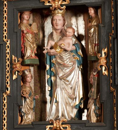 Zdjęcie nr 42: Retabulum ołtarzowe z dużą rzeźbą Matki Boskiej z Dzieciątkiem oraz mniejszymi o połowę czterema rzeźbami Świętych Dziewic umieszczonymi w prostopadłościennej szafie na rzucie trapezu (z ukośnie ustawionymi ściankami bocznymi). Rzeźby pełnoplastyczne, ścięte z tyłu, drążone, ustawione na cokolikach. Pośrodku figura Matki Boskiej w stylu gotyku międzynarodowego; zwrócona en face, w delikatnym kontrapoście, o esowato wygiętej sylwetce, podtrzymuje na obu rękach wpółleżące Dzieciatko. Twarz owalna z podwójnym podbródkiem, rysy twarzy płytko rzeźbione, delikatne, długi, prosty nos. Włosy upięte z tyłu. Maria ubrana w srebrzoną suknię z tkaniną drapowaną po środku w trzy fałdy misowe oraz złocony płaszcz podbity na niebieską podszewką, spływający od tyłu głowy na ramiona i boki, w układzie kaskadowym. Na głowie korona otwarta. Dzieciatko wpółleżące, zwrócone en trois quarts w prawo, prawą ręka ujmuje Matkę za szyję, w lewej trzyma złotą kulę. Twarz okrągła, duże uszy, włosy krótkie, kręcone. W partii bioder przepasany ściśle przylegającą, złotą tkaniną, delikatnie drapowaną. Na lewej ściance szafy u góry figurka św. Cecylii, zwróconej en face, o esowato wygiętej sylwetce, w lewej ręce, opartej na biodrze trzyma piszczałki, prawa wyciągnięta do przodu. Twarz okrągła o pełnych policzkach i płytko rzeźbionych rysach twarzy, włosy długie, falujące fakturowane, spływające przez ramiona na plecy. Ubrana w srebrzoną suknię oraz złocony płaszcz z czerwoną podszewką, spięty na piersi fibulą, z obfitą draperią przewieszoną przez ramiona, opadająca na froncie fałdami misowymi oraz spływającymi kaskadami od ramion. Na głowie blok drewna z namalowaną koroną otwartą.Na lewej ściance u dołu figura św. Katarzyny Aleksandryjskiej, zwróconej en face, w prawej ręce podtrzymuje koło, lewą wyciąga do przodu. Twarz okrągła, policzki pełne, o wysokim czole, nos duży, oczy duże delikatnie zarysowane. Włosy falowane, fakturowane, spływają na plecy. Święta ubrana w srebrzona suknię przepasaną w talii oraz złocony płaszcz spięty broszą na piersi, z obfitą draperią przewieszoną przez ramiona, opadająca na froncie misowymi fałdami i spływającymi kaskadami od ramion. Na głowie blok drewna z namalowaną koroną otwartą. Na prawej ściance u góry figura św. Doroty, zwrócona en face w delikatnym kontrapoście, prawa ręką podtrzymuje koszyk, lewą wskazuje na niego. Twarz owalna, pełna, delikatne rysy, wąski zadarty nos. Włosy falowane, fakturowane, opadają na tył głowy. Ubrana w złotą suknię, delikatnie drapowaną u dołu, oraz srebrzony płaszcz podbity czerwoną podszewką, spięty na piersi i układający się z przodu w fałdy misowe. Na głowie blok drewna z namalowaną koroną otwartą. Na prawej ściance u dołu figura św. Barbary, zwróconej en face, o esowato przegiętej sylwetce, obiema rękami przytrzymuje opartą na lewym ramieniu wieżę. Twarz owalna, pełna, delikatne rysy, wąski zadarty nos. Włosy falowane, fakturowane, opadają na tył głowy. Ubrana w suknię i srebrzony płaszcz z niebieską podszewką, spięty na piersi fibulą, przerzucony przez ramiona z obfitą partią materiału układającą się z przodu w fałdy misowe. Na głowie blok drewna z namalowaną koroną otwartą. Karnacje w tonacji naturalnej, szaty srebrzone i złocone.