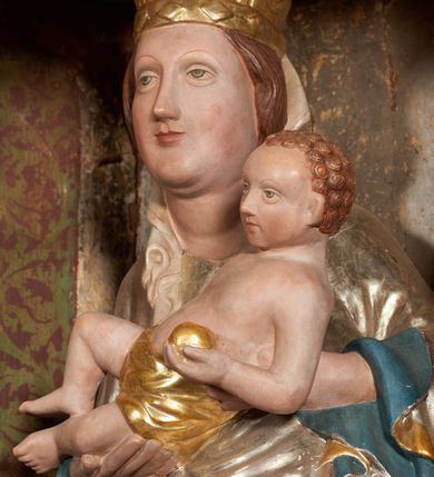 Zdjęcie nr 12: Retabulum ołtarzowe z dużą rzeźbą Matki Boskiej z Dzieciątkiem oraz mniejszymi o połowę czterema rzeźbami Świętych Dziewic umieszczonymi w prostopadłościennej szafie na rzucie trapezu (z ukośnie ustawionymi ściankami bocznymi). Rzeźby pełnoplastyczne, ścięte z tyłu, drążone, ustawione na cokolikach. Pośrodku figura Matki Boskiej w stylu gotyku międzynarodowego; zwrócona en face, w delikatnym kontrapoście, o esowato wygiętej sylwetce, podtrzymuje na obu rękach wpółleżące Dzieciatko. Twarz owalna z podwójnym podbródkiem, rysy twarzy płytko rzeźbione, delikatne, długi, prosty nos. Włosy upięte z tyłu. Maria ubrana w srebrzoną suknię z tkaniną drapowaną po środku w trzy fałdy misowe oraz złocony płaszcz podbity na niebieską podszewką, spływający od tyłu głowy na ramiona i boki, w układzie kaskadowym. Na głowie korona otwarta. Dzieciatko wpółleżące, zwrócone en trois quarts w prawo, prawą ręka ujmuje Matkę za szyję, w lewej trzyma złotą kulę. Twarz okrągła, duże uszy, włosy krótkie, kręcone. W partii bioder przepasany ściśle przylegającą, złotą tkaniną, delikatnie drapowaną. Na lewej ściance szafy u góry figurka św. Cecylii, zwróconej en face, o esowato wygiętej sylwetce, w lewej ręce, opartej na biodrze trzyma piszczałki, prawa wyciągnięta do przodu. Twarz okrągła o pełnych policzkach i płytko rzeźbionych rysach twarzy, włosy długie, falujące fakturowane, spływające przez ramiona na plecy. Ubrana w srebrzoną suknię oraz złocony płaszcz z czerwoną podszewką, spięty na piersi fibulą, z obfitą draperią przewieszoną przez ramiona, opadająca na froncie fałdami misowymi oraz spływającymi kaskadami od ramion. Na głowie blok drewna z namalowaną koroną otwartą.Na lewej ściance u dołu figura św. Katarzyny Aleksandryjskiej, zwróconej en face, w prawej ręce podtrzymuje koło, lewą wyciąga do przodu. Twarz okrągła, policzki pełne, o wysokim czole, nos duży, oczy duże delikatnie zarysowane. Włosy falowane, fakturowane, spływają na plecy. Święta ubrana w srebrzona suknię przepasaną w talii oraz złocony płaszcz spięty broszą na piersi, z obfitą draperią przewieszoną przez ramiona, opadająca na froncie misowymi fałdami i spływającymi kaskadami od ramion. Na głowie blok drewna z namalowaną koroną otwartą. Na prawej ściance u góry figura św. Doroty, zwrócona en face w delikatnym kontrapoście, prawa ręką podtrzymuje koszyk, lewą wskazuje na niego. Twarz owalna, pełna, delikatne rysy, wąski zadarty nos. Włosy falowane, fakturowane, opadają na tył głowy. Ubrana w złotą suknię, delikatnie drapowaną u dołu, oraz srebrzony płaszcz podbity czerwoną podszewką, spięty na piersi i układający się z przodu w fałdy misowe. Na głowie blok drewna z namalowaną koroną otwartą. Na prawej ściance u dołu figura św. Barbary, zwróconej en face, o esowato przegiętej sylwetce, obiema rękami przytrzymuje opartą na lewym ramieniu wieżę. Twarz owalna, pełna, delikatne rysy, wąski zadarty nos. Włosy falowane, fakturowane, opadają na tył głowy. Ubrana w suknię i srebrzony płaszcz z niebieską podszewką, spięty na piersi fibulą, przerzucony przez ramiona z obfitą partią materiału układającą się z przodu w fałdy misowe. Na głowie blok drewna z namalowaną koroną otwartą. Karnacje w tonacji naturalnej, szaty srebrzone i złocone.