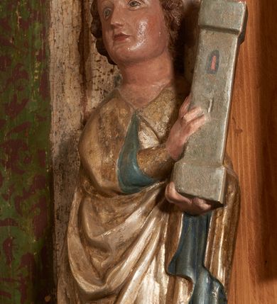 Zdjęcie nr 31: Retabulum ołtarzowe z dużą rzeźbą Matki Boskiej z Dzieciątkiem oraz mniejszymi o połowę czterema rzeźbami Świętych Dziewic umieszczonymi w prostopadłościennej szafie na rzucie trapezu (z ukośnie ustawionymi ściankami bocznymi). Rzeźby pełnoplastyczne, ścięte z tyłu, drążone, ustawione na cokolikach. Pośrodku figura Matki Boskiej w stylu gotyku międzynarodowego; zwrócona en face, w delikatnym kontrapoście, o esowato wygiętej sylwetce, podtrzymuje na obu rękach wpółleżące Dzieciatko. Twarz owalna z podwójnym podbródkiem, rysy twarzy płytko rzeźbione, delikatne, długi, prosty nos. Włosy upięte z tyłu. Maria ubrana w srebrzoną suknię z tkaniną drapowaną po środku w trzy fałdy misowe oraz złocony płaszcz podbity na niebieską podszewką, spływający od tyłu głowy na ramiona i boki, w układzie kaskadowym. Na głowie korona otwarta. Dzieciatko wpółleżące, zwrócone en trois quarts w prawo, prawą ręka ujmuje Matkę za szyję, w lewej trzyma złotą kulę. Twarz okrągła, duże uszy, włosy krótkie, kręcone. W partii bioder przepasany ściśle przylegającą, złotą tkaniną, delikatnie drapowaną. Na lewej ściance szafy u góry figurka św. Cecylii, zwróconej en face, o esowato wygiętej sylwetce, w lewej ręce, opartej na biodrze trzyma piszczałki, prawa wyciągnięta do przodu. Twarz okrągła o pełnych policzkach i płytko rzeźbionych rysach twarzy, włosy długie, falujące fakturowane, spływające przez ramiona na plecy. Ubrana w srebrzoną suknię oraz złocony płaszcz z czerwoną podszewką, spięty na piersi fibulą, z obfitą draperią przewieszoną przez ramiona, opadająca na froncie fałdami misowymi oraz spływającymi kaskadami od ramion. Na głowie blok drewna z namalowaną koroną otwartą.Na lewej ściance u dołu figura św. Katarzyny Aleksandryjskiej, zwróconej en face, w prawej ręce podtrzymuje koło, lewą wyciąga do przodu. Twarz okrągła, policzki pełne, o wysokim czole, nos duży, oczy duże delikatnie zarysowane. Włosy falowane, fakturowane, spływają na plecy. Święta ubrana w srebrzona suknię przepasaną w talii oraz złocony płaszcz spięty broszą na piersi, z obfitą draperią przewieszoną przez ramiona, opadająca na froncie misowymi fałdami i spływającymi kaskadami od ramion. Na głowie blok drewna z namalowaną koroną otwartą. Na prawej ściance u góry figura św. Doroty, zwrócona en face w delikatnym kontrapoście, prawa ręką podtrzymuje koszyk, lewą wskazuje na niego. Twarz owalna, pełna, delikatne rysy, wąski zadarty nos. Włosy falowane, fakturowane, opadają na tył głowy. Ubrana w złotą suknię, delikatnie drapowaną u dołu, oraz srebrzony płaszcz podbity czerwoną podszewką, spięty na piersi i układający się z przodu w fałdy misowe. Na głowie blok drewna z namalowaną koroną otwartą. Na prawej ściance u dołu figura św. Barbary, zwróconej en face, o esowato przegiętej sylwetce, obiema rękami przytrzymuje opartą na lewym ramieniu wieżę. Twarz owalna, pełna, delikatne rysy, wąski zadarty nos. Włosy falowane, fakturowane, opadają na tył głowy. Ubrana w suknię i srebrzony płaszcz z niebieską podszewką, spięty na piersi fibulą, przerzucony przez ramiona z obfitą partią materiału układającą się z przodu w fałdy misowe. Na głowie blok drewna z namalowaną koroną otwartą. Karnacje w tonacji naturalnej, szaty srebrzone i złocone.