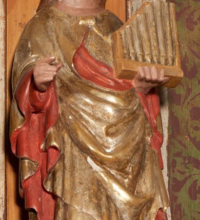 Zdjęcie nr 20: Retabulum ołtarzowe z dużą rzeźbą Matki Boskiej z Dzieciątkiem oraz mniejszymi o połowę czterema rzeźbami Świętych Dziewic umieszczonymi w prostopadłościennej szafie na rzucie trapezu (z ukośnie ustawionymi ściankami bocznymi). Rzeźby pełnoplastyczne, ścięte z tyłu, drążone, ustawione na cokolikach. Pośrodku figura Matki Boskiej w stylu gotyku międzynarodowego; zwrócona en face, w delikatnym kontrapoście, o esowato wygiętej sylwetce, podtrzymuje na obu rękach wpółleżące Dzieciatko. Twarz owalna z podwójnym podbródkiem, rysy twarzy płytko rzeźbione, delikatne, długi, prosty nos. Włosy upięte z tyłu. Maria ubrana w srebrzoną suknię z tkaniną drapowaną po środku w trzy fałdy misowe oraz złocony płaszcz podbity na niebieską podszewką, spływający od tyłu głowy na ramiona i boki, w układzie kaskadowym. Na głowie korona otwarta. Dzieciatko wpółleżące, zwrócone en trois quarts w prawo, prawą ręka ujmuje Matkę za szyję, w lewej trzyma złotą kulę. Twarz okrągła, duże uszy, włosy krótkie, kręcone. W partii bioder przepasany ściśle przylegającą, złotą tkaniną, delikatnie drapowaną. Na lewej ściance szafy u góry figurka św. Cecylii, zwróconej en face, o esowato wygiętej sylwetce, w lewej ręce, opartej na biodrze trzyma piszczałki, prawa wyciągnięta do przodu. Twarz okrągła o pełnych policzkach i płytko rzeźbionych rysach twarzy, włosy długie, falujące fakturowane, spływające przez ramiona na plecy. Ubrana w srebrzoną suknię oraz złocony płaszcz z czerwoną podszewką, spięty na piersi fibulą, z obfitą draperią przewieszoną przez ramiona, opadająca na froncie fałdami misowymi oraz spływającymi kaskadami od ramion. Na głowie blok drewna z namalowaną koroną otwartą.Na lewej ściance u dołu figura św. Katarzyny Aleksandryjskiej, zwróconej en face, w prawej ręce podtrzymuje koło, lewą wyciąga do przodu. Twarz okrągła, policzki pełne, o wysokim czole, nos duży, oczy duże delikatnie zarysowane. Włosy falowane, fakturowane, spływają na plecy. Święta ubrana w srebrzona suknię przepasaną w talii oraz złocony płaszcz spięty broszą na piersi, z obfitą draperią przewieszoną przez ramiona, opadająca na froncie misowymi fałdami i spływającymi kaskadami od ramion. Na głowie blok drewna z namalowaną koroną otwartą. Na prawej ściance u góry figura św. Doroty, zwrócona en face w delikatnym kontrapoście, prawa ręką podtrzymuje koszyk, lewą wskazuje na niego. Twarz owalna, pełna, delikatne rysy, wąski zadarty nos. Włosy falowane, fakturowane, opadają na tył głowy. Ubrana w złotą suknię, delikatnie drapowaną u dołu, oraz srebrzony płaszcz podbity czerwoną podszewką, spięty na piersi i układający się z przodu w fałdy misowe. Na głowie blok drewna z namalowaną koroną otwartą. Na prawej ściance u dołu figura św. Barbary, zwróconej en face, o esowato przegiętej sylwetce, obiema rękami przytrzymuje opartą na lewym ramieniu wieżę. Twarz owalna, pełna, delikatne rysy, wąski zadarty nos. Włosy falowane, fakturowane, opadają na tył głowy. Ubrana w suknię i srebrzony płaszcz z niebieską podszewką, spięty na piersi fibulą, przerzucony przez ramiona z obfitą partią materiału układającą się z przodu w fałdy misowe. Na głowie blok drewna z namalowaną koroną otwartą. Karnacje w tonacji naturalnej, szaty srebrzone i złocone.