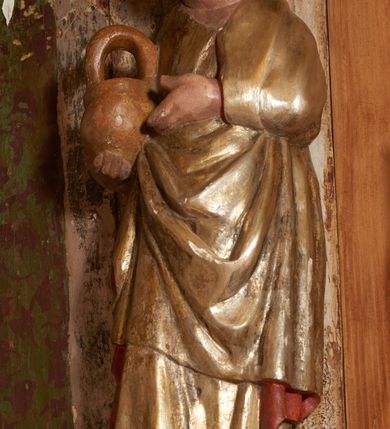 Zdjęcie nr 28: Retabulum ołtarzowe z dużą rzeźbą Matki Boskiej z Dzieciątkiem oraz mniejszymi o połowę czterema rzeźbami Świętych Dziewic umieszczonymi w prostopadłościennej szafie na rzucie trapezu (z ukośnie ustawionymi ściankami bocznymi). Rzeźby pełnoplastyczne, ścięte z tyłu, drążone, ustawione na cokolikach. Pośrodku figura Matki Boskiej w stylu gotyku międzynarodowego; zwrócona en face, w delikatnym kontrapoście, o esowato wygiętej sylwetce, podtrzymuje na obu rękach wpółleżące Dzieciatko. Twarz owalna z podwójnym podbródkiem, rysy twarzy płytko rzeźbione, delikatne, długi, prosty nos. Włosy upięte z tyłu. Maria ubrana w srebrzoną suknię z tkaniną drapowaną po środku w trzy fałdy misowe oraz złocony płaszcz podbity na niebieską podszewką, spływający od tyłu głowy na ramiona i boki, w układzie kaskadowym. Na głowie korona otwarta. Dzieciatko wpółleżące, zwrócone en trois quarts w prawo, prawą ręka ujmuje Matkę za szyję, w lewej trzyma złotą kulę. Twarz okrągła, duże uszy, włosy krótkie, kręcone. W partii bioder przepasany ściśle przylegającą, złotą tkaniną, delikatnie drapowaną. Na lewej ściance szafy u góry figurka św. Cecylii, zwróconej en face, o esowato wygiętej sylwetce, w lewej ręce, opartej na biodrze trzyma piszczałki, prawa wyciągnięta do przodu. Twarz okrągła o pełnych policzkach i płytko rzeźbionych rysach twarzy, włosy długie, falujące fakturowane, spływające przez ramiona na plecy. Ubrana w srebrzoną suknię oraz złocony płaszcz z czerwoną podszewką, spięty na piersi fibulą, z obfitą draperią przewieszoną przez ramiona, opadająca na froncie fałdami misowymi oraz spływającymi kaskadami od ramion. Na głowie blok drewna z namalowaną koroną otwartą.Na lewej ściance u dołu figura św. Katarzyny Aleksandryjskiej, zwróconej en face, w prawej ręce podtrzymuje koło, lewą wyciąga do przodu. Twarz okrągła, policzki pełne, o wysokim czole, nos duży, oczy duże delikatnie zarysowane. Włosy falowane, fakturowane, spływają na plecy. Święta ubrana w srebrzona suknię przepasaną w talii oraz złocony płaszcz spięty broszą na piersi, z obfitą draperią przewieszoną przez ramiona, opadająca na froncie misowymi fałdami i spływającymi kaskadami od ramion. Na głowie blok drewna z namalowaną koroną otwartą. Na prawej ściance u góry figura św. Doroty, zwrócona en face w delikatnym kontrapoście, prawa ręką podtrzymuje koszyk, lewą wskazuje na niego. Twarz owalna, pełna, delikatne rysy, wąski zadarty nos. Włosy falowane, fakturowane, opadają na tył głowy. Ubrana w złotą suknię, delikatnie drapowaną u dołu, oraz srebrzony płaszcz podbity czerwoną podszewką, spięty na piersi i układający się z przodu w fałdy misowe. Na głowie blok drewna z namalowaną koroną otwartą. Na prawej ściance u dołu figura św. Barbary, zwróconej en face, o esowato przegiętej sylwetce, obiema rękami przytrzymuje opartą na lewym ramieniu wieżę. Twarz owalna, pełna, delikatne rysy, wąski zadarty nos. Włosy falowane, fakturowane, opadają na tył głowy. Ubrana w suknię i srebrzony płaszcz z niebieską podszewką, spięty na piersi fibulą, przerzucony przez ramiona z obfitą partią materiału układającą się z przodu w fałdy misowe. Na głowie blok drewna z namalowaną koroną otwartą. Karnacje w tonacji naturalnej, szaty srebrzone i złocone.