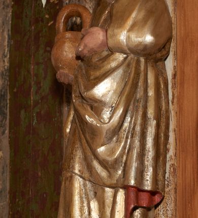 Zdjęcie nr 29: Retabulum ołtarzowe z dużą rzeźbą Matki Boskiej z Dzieciątkiem oraz mniejszymi o połowę czterema rzeźbami Świętych Dziewic umieszczonymi w prostopadłościennej szafie na rzucie trapezu (z ukośnie ustawionymi ściankami bocznymi). Rzeźby pełnoplastyczne, ścięte z tyłu, drążone, ustawione na cokolikach. Pośrodku figura Matki Boskiej w stylu gotyku międzynarodowego; zwrócona en face, w delikatnym kontrapoście, o esowato wygiętej sylwetce, podtrzymuje na obu rękach wpółleżące Dzieciatko. Twarz owalna z podwójnym podbródkiem, rysy twarzy płytko rzeźbione, delikatne, długi, prosty nos. Włosy upięte z tyłu. Maria ubrana w srebrzoną suknię z tkaniną drapowaną po środku w trzy fałdy misowe oraz złocony płaszcz podbity na niebieską podszewką, spływający od tyłu głowy na ramiona i boki, w układzie kaskadowym. Na głowie korona otwarta. Dzieciatko wpółleżące, zwrócone en trois quarts w prawo, prawą ręka ujmuje Matkę za szyję, w lewej trzyma złotą kulę. Twarz okrągła, duże uszy, włosy krótkie, kręcone. W partii bioder przepasany ściśle przylegającą, złotą tkaniną, delikatnie drapowaną. Na lewej ściance szafy u góry figurka św. Cecylii, zwróconej en face, o esowato wygiętej sylwetce, w lewej ręce, opartej na biodrze trzyma piszczałki, prawa wyciągnięta do przodu. Twarz okrągła o pełnych policzkach i płytko rzeźbionych rysach twarzy, włosy długie, falujące fakturowane, spływające przez ramiona na plecy. Ubrana w srebrzoną suknię oraz złocony płaszcz z czerwoną podszewką, spięty na piersi fibulą, z obfitą draperią przewieszoną przez ramiona, opadająca na froncie fałdami misowymi oraz spływającymi kaskadami od ramion. Na głowie blok drewna z namalowaną koroną otwartą.Na lewej ściance u dołu figura św. Katarzyny Aleksandryjskiej, zwróconej en face, w prawej ręce podtrzymuje koło, lewą wyciąga do przodu. Twarz okrągła, policzki pełne, o wysokim czole, nos duży, oczy duże delikatnie zarysowane. Włosy falowane, fakturowane, spływają na plecy. Święta ubrana w srebrzona suknię przepasaną w talii oraz złocony płaszcz spięty broszą na piersi, z obfitą draperią przewieszoną przez ramiona, opadająca na froncie misowymi fałdami i spływającymi kaskadami od ramion. Na głowie blok drewna z namalowaną koroną otwartą. Na prawej ściance u góry figura św. Doroty, zwrócona en face w delikatnym kontrapoście, prawa ręką podtrzymuje koszyk, lewą wskazuje na niego. Twarz owalna, pełna, delikatne rysy, wąski zadarty nos. Włosy falowane, fakturowane, opadają na tył głowy. Ubrana w złotą suknię, delikatnie drapowaną u dołu, oraz srebrzony płaszcz podbity czerwoną podszewką, spięty na piersi i układający się z przodu w fałdy misowe. Na głowie blok drewna z namalowaną koroną otwartą. Na prawej ściance u dołu figura św. Barbary, zwróconej en face, o esowato przegiętej sylwetce, obiema rękami przytrzymuje opartą na lewym ramieniu wieżę. Twarz owalna, pełna, delikatne rysy, wąski zadarty nos. Włosy falowane, fakturowane, opadają na tył głowy. Ubrana w suknię i srebrzony płaszcz z niebieską podszewką, spięty na piersi fibulą, przerzucony przez ramiona z obfitą partią materiału układającą się z przodu w fałdy misowe. Na głowie blok drewna z namalowaną koroną otwartą. Karnacje w tonacji naturalnej, szaty srebrzone i złocone.