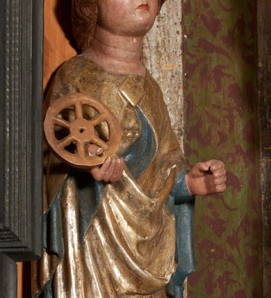 Zdjęcie nr 25: Retabulum ołtarzowe z dużą rzeźbą Matki Boskiej z Dzieciątkiem oraz mniejszymi o połowę czterema rzeźbami Świętych Dziewic umieszczonymi w prostopadłościennej szafie na rzucie trapezu (z ukośnie ustawionymi ściankami bocznymi). Rzeźby pełnoplastyczne, ścięte z tyłu, drążone, ustawione na cokolikach. Pośrodku figura Matki Boskiej w stylu gotyku międzynarodowego; zwrócona en face, w delikatnym kontrapoście, o esowato wygiętej sylwetce, podtrzymuje na obu rękach wpółleżące Dzieciatko. Twarz owalna z podwójnym podbródkiem, rysy twarzy płytko rzeźbione, delikatne, długi, prosty nos. Włosy upięte z tyłu. Maria ubrana w srebrzoną suknię z tkaniną drapowaną po środku w trzy fałdy misowe oraz złocony płaszcz podbity na niebieską podszewką, spływający od tyłu głowy na ramiona i boki, w układzie kaskadowym. Na głowie korona otwarta. Dzieciatko wpółleżące, zwrócone en trois quarts w prawo, prawą ręka ujmuje Matkę za szyję, w lewej trzyma złotą kulę. Twarz okrągła, duże uszy, włosy krótkie, kręcone. W partii bioder przepasany ściśle przylegającą, złotą tkaniną, delikatnie drapowaną. Na lewej ściance szafy u góry figurka św. Cecylii, zwróconej en face, o esowato wygiętej sylwetce, w lewej ręce, opartej na biodrze trzyma piszczałki, prawa wyciągnięta do przodu. Twarz okrągła o pełnych policzkach i płytko rzeźbionych rysach twarzy, włosy długie, falujące fakturowane, spływające przez ramiona na plecy. Ubrana w srebrzoną suknię oraz złocony płaszcz z czerwoną podszewką, spięty na piersi fibulą, z obfitą draperią przewieszoną przez ramiona, opadająca na froncie fałdami misowymi oraz spływającymi kaskadami od ramion. Na głowie blok drewna z namalowaną koroną otwartą.Na lewej ściance u dołu figura św. Katarzyny Aleksandryjskiej, zwróconej en face, w prawej ręce podtrzymuje koło, lewą wyciąga do przodu. Twarz okrągła, policzki pełne, o wysokim czole, nos duży, oczy duże delikatnie zarysowane. Włosy falowane, fakturowane, spływają na plecy. Święta ubrana w srebrzona suknię przepasaną w talii oraz złocony płaszcz spięty broszą na piersi, z obfitą draperią przewieszoną przez ramiona, opadająca na froncie misowymi fałdami i spływającymi kaskadami od ramion. Na głowie blok drewna z namalowaną koroną otwartą. Na prawej ściance u góry figura św. Doroty, zwrócona en face w delikatnym kontrapoście, prawa ręką podtrzymuje koszyk, lewą wskazuje na niego. Twarz owalna, pełna, delikatne rysy, wąski zadarty nos. Włosy falowane, fakturowane, opadają na tył głowy. Ubrana w złotą suknię, delikatnie drapowaną u dołu, oraz srebrzony płaszcz podbity czerwoną podszewką, spięty na piersi i układający się z przodu w fałdy misowe. Na głowie blok drewna z namalowaną koroną otwartą. Na prawej ściance u dołu figura św. Barbary, zwróconej en face, o esowato przegiętej sylwetce, obiema rękami przytrzymuje opartą na lewym ramieniu wieżę. Twarz owalna, pełna, delikatne rysy, wąski zadarty nos. Włosy falowane, fakturowane, opadają na tył głowy. Ubrana w suknię i srebrzony płaszcz z niebieską podszewką, spięty na piersi fibulą, przerzucony przez ramiona z obfitą partią materiału układającą się z przodu w fałdy misowe. Na głowie blok drewna z namalowaną koroną otwartą. Karnacje w tonacji naturalnej, szaty srebrzone i złocone.