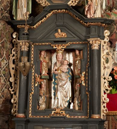 Zdjęcie nr 1: Retabulum ołtarzowe z dużą rzeźbą Matki Boskiej z Dzieciątkiem oraz mniejszymi o połowę czterema rzeźbami Świętych Dziewic umieszczonymi w prostopadłościennej szafie na rzucie trapezu (z ukośnie ustawionymi ściankami bocznymi). Rzeźby pełnoplastyczne, ścięte z tyłu, drążone, ustawione na cokolikach. Pośrodku figura Matki Boskiej w stylu gotyku międzynarodowego; zwrócona en face, w delikatnym kontrapoście, o esowato wygiętej sylwetce, podtrzymuje na obu rękach wpółleżące Dzieciatko. Twarz owalna z podwójnym podbródkiem, rysy twarzy płytko rzeźbione, delikatne, długi, prosty nos. Włosy upięte z tyłu. Maria ubrana w srebrzoną suknię z tkaniną drapowaną po środku w trzy fałdy misowe oraz złocony płaszcz podbity na niebieską podszewką, spływający od tyłu głowy na ramiona i boki, w układzie kaskadowym. Na głowie korona otwarta. Dzieciatko wpółleżące, zwrócone en trois quarts w prawo, prawą ręka ujmuje Matkę za szyję, w lewej trzyma złotą kulę. Twarz okrągła, duże uszy, włosy krótkie, kręcone. W partii bioder przepasany ściśle przylegającą, złotą tkaniną, delikatnie drapowaną. Na lewej ściance szafy u góry figurka św. Cecylii, zwróconej en face, o esowato wygiętej sylwetce, w lewej ręce, opartej na biodrze trzyma piszczałki, prawa wyciągnięta do przodu. Twarz okrągła o pełnych policzkach i płytko rzeźbionych rysach twarzy, włosy długie, falujące fakturowane, spływające przez ramiona na plecy. Ubrana w srebrzoną suknię oraz złocony płaszcz z czerwoną podszewką, spięty na piersi fibulą, z obfitą draperią przewieszoną przez ramiona, opadająca na froncie fałdami misowymi oraz spływającymi kaskadami od ramion. Na głowie blok drewna z namalowaną koroną otwartą.Na lewej ściance u dołu figura św. Katarzyny Aleksandryjskiej, zwróconej en face, w prawej ręce podtrzymuje koło, lewą wyciąga do przodu. Twarz okrągła, policzki pełne, o wysokim czole, nos duży, oczy duże delikatnie zarysowane. Włosy falowane, fakturowane, spływają na plecy. Święta ubrana w srebrzona suknię przepasaną w talii oraz złocony płaszcz spięty broszą na piersi, z obfitą draperią przewieszoną przez ramiona, opadająca na froncie misowymi fałdami i spływającymi kaskadami od ramion. Na głowie blok drewna z namalowaną koroną otwartą. Na prawej ściance u góry figura św. Doroty, zwrócona en face w delikatnym kontrapoście, prawa ręką podtrzymuje koszyk, lewą wskazuje na niego. Twarz owalna, pełna, delikatne rysy, wąski zadarty nos. Włosy falowane, fakturowane, opadają na tył głowy. Ubrana w złotą suknię, delikatnie drapowaną u dołu, oraz srebrzony płaszcz podbity czerwoną podszewką, spięty na piersi i układający się z przodu w fałdy misowe. Na głowie blok drewna z namalowaną koroną otwartą. Na prawej ściance u dołu figura św. Barbary, zwróconej en face, o esowato przegiętej sylwetce, obiema rękami przytrzymuje opartą na lewym ramieniu wieżę. Twarz owalna, pełna, delikatne rysy, wąski zadarty nos. Włosy falowane, fakturowane, opadają na tył głowy. Ubrana w suknię i srebrzony płaszcz z niebieską podszewką, spięty na piersi fibulą, przerzucony przez ramiona z obfitą partią materiału układającą się z przodu w fałdy misowe. Na głowie blok drewna z namalowaną koroną otwartą. Karnacje w tonacji naturalnej, szaty srebrzone i złocone.