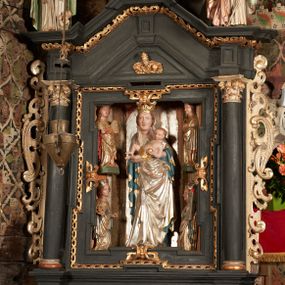 Zdjęcie nr 1: Retabulum ołtarzowe z dużą rzeźbą Matki Boskiej z Dzieciątkiem oraz mniejszymi o połowę czterema rzeźbami Świętych Dziewic umieszczonymi w prostopadłościennej szafie na rzucie trapezu (z ukośnie ustawionymi ściankami bocznymi). Rzeźby pełnoplastyczne, ścięte z tyłu, drążone, ustawione na cokolikach. Pośrodku figura Matki Boskiej w stylu gotyku międzynarodowego; zwrócona en face, w delikatnym kontrapoście, o esowato wygiętej sylwetce, podtrzymuje na obu rękach wpółleżące Dzieciatko. Twarz owalna z podwójnym podbródkiem, rysy twarzy płytko rzeźbione, delikatne, długi, prosty nos. Włosy upięte z tyłu. Maria ubrana w srebrzoną suknię z tkaniną drapowaną po środku w trzy fałdy misowe oraz złocony płaszcz podbity na niebieską podszewką, spływający od tyłu głowy na ramiona i boki, w układzie kaskadowym. Na głowie korona otwarta. Dzieciatko wpółleżące, zwrócone en trois quarts w prawo, prawą ręka ujmuje Matkę za szyję, w lewej trzyma złotą kulę. Twarz okrągła, duże uszy, włosy krótkie, kręcone. W partii bioder przepasany ściśle przylegającą, złotą tkaniną, delikatnie drapowaną. Na lewej ściance szafy u góry figurka św. Cecylii, zwróconej en face, o esowato wygiętej sylwetce, w lewej ręce, opartej na biodrze trzyma piszczałki, prawa wyciągnięta do przodu. Twarz okrągła o pełnych policzkach i płytko rzeźbionych rysach twarzy, włosy długie, falujące fakturowane, spływające przez ramiona na plecy. Ubrana w srebrzoną suknię oraz złocony płaszcz z czerwoną podszewką, spięty na piersi fibulą, z obfitą draperią przewieszoną przez ramiona, opadająca na froncie fałdami misowymi oraz spływającymi kaskadami od ramion. Na głowie blok drewna z namalowaną koroną otwartą.Na lewej ściance u dołu figura św. Katarzyny Aleksandryjskiej, zwróconej en face, w prawej ręce podtrzymuje koło, lewą wyciąga do przodu. Twarz okrągła, policzki pełne, o wysokim czole, nos duży, oczy duże delikatnie zarysowane. Włosy falowane, fakturowane, spływają na plecy. Święta ubrana w srebrzona suknię przepasaną w talii oraz złocony płaszcz spięty broszą na piersi, z obfitą draperią przewieszoną przez ramiona, opadająca na froncie misowymi fałdami i spływającymi kaskadami od ramion. Na głowie blok drewna z namalowaną koroną otwartą. Na prawej ściance u góry figura św. Doroty, zwrócona en face w delikatnym kontrapoście, prawa ręką podtrzymuje koszyk, lewą wskazuje na niego. Twarz owalna, pełna, delikatne rysy, wąski zadarty nos. Włosy falowane, fakturowane, opadają na tył głowy. Ubrana w złotą suknię, delikatnie drapowaną u dołu, oraz srebrzony płaszcz podbity czerwoną podszewką, spięty na piersi i układający się z przodu w fałdy misowe. Na głowie blok drewna z namalowaną koroną otwartą. Na prawej ściance u dołu figura św. Barbary, zwróconej en face, o esowato przegiętej sylwetce, obiema rękami przytrzymuje opartą na lewym ramieniu wieżę. Twarz owalna, pełna, delikatne rysy, wąski zadarty nos. Włosy falowane, fakturowane, opadają na tył głowy. Ubrana w suknię i srebrzony płaszcz z niebieską podszewką, spięty na piersi fibulą, przerzucony przez ramiona z obfitą partią materiału układającą się z przodu w fałdy misowe. Na głowie blok drewna z namalowaną koroną otwartą. Karnacje w tonacji naturalnej, szaty srebrzone i złocone.