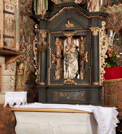 Zdjęcie nr 2: Retabulum ołtarzowe z dużą rzeźbą Matki Boskiej z Dzieciątkiem oraz mniejszymi o połowę czterema rzeźbami Świętych Dziewic umieszczonymi w prostopadłościennej szafie na rzucie trapezu (z ukośnie ustawionymi ściankami bocznymi). Rzeźby pełnoplastyczne, ścięte z tyłu, drążone, ustawione na cokolikach. Pośrodku figura Matki Boskiej w stylu gotyku międzynarodowego; zwrócona en face, w delikatnym kontrapoście, o esowato wygiętej sylwetce, podtrzymuje na obu rękach wpółleżące Dzieciatko. Twarz owalna z podwójnym podbródkiem, rysy twarzy płytko rzeźbione, delikatne, długi, prosty nos. Włosy upięte z tyłu. Maria ubrana w srebrzoną suknię z tkaniną drapowaną po środku w trzy fałdy misowe oraz złocony płaszcz podbity na niebieską podszewką, spływający od tyłu głowy na ramiona i boki, w układzie kaskadowym. Na głowie korona otwarta. Dzieciatko wpółleżące, zwrócone en trois quarts w prawo, prawą ręka ujmuje Matkę za szyję, w lewej trzyma złotą kulę. Twarz okrągła, duże uszy, włosy krótkie, kręcone. W partii bioder przepasany ściśle przylegającą, złotą tkaniną, delikatnie drapowaną. Na lewej ściance szafy u góry figurka św. Cecylii, zwróconej en face, o esowato wygiętej sylwetce, w lewej ręce, opartej na biodrze trzyma piszczałki, prawa wyciągnięta do przodu. Twarz okrągła o pełnych policzkach i płytko rzeźbionych rysach twarzy, włosy długie, falujące fakturowane, spływające przez ramiona na plecy. Ubrana w srebrzoną suknię oraz złocony płaszcz z czerwoną podszewką, spięty na piersi fibulą, z obfitą draperią przewieszoną przez ramiona, opadająca na froncie fałdami misowymi oraz spływającymi kaskadami od ramion. Na głowie blok drewna z namalowaną koroną otwartą.Na lewej ściance u dołu figura św. Katarzyny Aleksandryjskiej, zwróconej en face, w prawej ręce podtrzymuje koło, lewą wyciąga do przodu. Twarz okrągła, policzki pełne, o wysokim czole, nos duży, oczy duże delikatnie zarysowane. Włosy falowane, fakturowane, spływają na plecy. Święta ubrana w srebrzona suknię przepasaną w talii oraz złocony płaszcz spięty broszą na piersi, z obfitą draperią przewieszoną przez ramiona, opadająca na froncie misowymi fałdami i spływającymi kaskadami od ramion. Na głowie blok drewna z namalowaną koroną otwartą. Na prawej ściance u góry figura św. Doroty, zwrócona en face w delikatnym kontrapoście, prawa ręką podtrzymuje koszyk, lewą wskazuje na niego. Twarz owalna, pełna, delikatne rysy, wąski zadarty nos. Włosy falowane, fakturowane, opadają na tył głowy. Ubrana w złotą suknię, delikatnie drapowaną u dołu, oraz srebrzony płaszcz podbity czerwoną podszewką, spięty na piersi i układający się z przodu w fałdy misowe. Na głowie blok drewna z namalowaną koroną otwartą. Na prawej ściance u dołu figura św. Barbary, zwróconej en face, o esowato przegiętej sylwetce, obiema rękami przytrzymuje opartą na lewym ramieniu wieżę. Twarz owalna, pełna, delikatne rysy, wąski zadarty nos. Włosy falowane, fakturowane, opadają na tył głowy. Ubrana w suknię i srebrzony płaszcz z niebieską podszewką, spięty na piersi fibulą, przerzucony przez ramiona z obfitą partią materiału układającą się z przodu w fałdy misowe. Na głowie blok drewna z namalowaną koroną otwartą. Karnacje w tonacji naturalnej, szaty srebrzone i złocone.