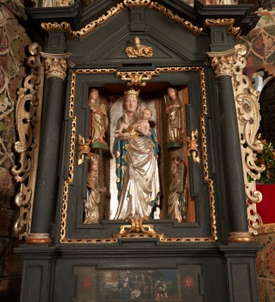 Zdjęcie nr 13: Retabulum ołtarzowe z dużą rzeźbą Matki Boskiej z Dzieciątkiem oraz mniejszymi o połowę czterema rzeźbami Świętych Dziewic umieszczonymi w prostopadłościennej szafie na rzucie trapezu (z ukośnie ustawionymi ściankami bocznymi). Rzeźby pełnoplastyczne, ścięte z tyłu, drążone, ustawione na cokolikach. Pośrodku figura Matki Boskiej w stylu gotyku międzynarodowego; zwrócona en face, w delikatnym kontrapoście, o esowato wygiętej sylwetce, podtrzymuje na obu rękach wpółleżące Dzieciatko. Twarz owalna z podwójnym podbródkiem, rysy twarzy płytko rzeźbione, delikatne, długi, prosty nos. Włosy upięte z tyłu. Maria ubrana w srebrzoną suknię z tkaniną drapowaną po środku w trzy fałdy misowe oraz złocony płaszcz podbity na niebieską podszewką, spływający od tyłu głowy na ramiona i boki, w układzie kaskadowym. Na głowie korona otwarta. Dzieciatko wpółleżące, zwrócone en trois quarts w prawo, prawą ręka ujmuje Matkę za szyję, w lewej trzyma złotą kulę. Twarz okrągła, duże uszy, włosy krótkie, kręcone. W partii bioder przepasany ściśle przylegającą, złotą tkaniną, delikatnie drapowaną. Na lewej ściance szafy u góry figurka św. Cecylii, zwróconej en face, o esowato wygiętej sylwetce, w lewej ręce, opartej na biodrze trzyma piszczałki, prawa wyciągnięta do przodu. Twarz okrągła o pełnych policzkach i płytko rzeźbionych rysach twarzy, włosy długie, falujące fakturowane, spływające przez ramiona na plecy. Ubrana w srebrzoną suknię oraz złocony płaszcz z czerwoną podszewką, spięty na piersi fibulą, z obfitą draperią przewieszoną przez ramiona, opadająca na froncie fałdami misowymi oraz spływającymi kaskadami od ramion. Na głowie blok drewna z namalowaną koroną otwartą.Na lewej ściance u dołu figura św. Katarzyny Aleksandryjskiej, zwróconej en face, w prawej ręce podtrzymuje koło, lewą wyciąga do przodu. Twarz okrągła, policzki pełne, o wysokim czole, nos duży, oczy duże delikatnie zarysowane. Włosy falowane, fakturowane, spływają na plecy. Święta ubrana w srebrzona suknię przepasaną w talii oraz złocony płaszcz spięty broszą na piersi, z obfitą draperią przewieszoną przez ramiona, opadająca na froncie misowymi fałdami i spływającymi kaskadami od ramion. Na głowie blok drewna z namalowaną koroną otwartą. Na prawej ściance u góry figura św. Doroty, zwrócona en face w delikatnym kontrapoście, prawa ręką podtrzymuje koszyk, lewą wskazuje na niego. Twarz owalna, pełna, delikatne rysy, wąski zadarty nos. Włosy falowane, fakturowane, opadają na tył głowy. Ubrana w złotą suknię, delikatnie drapowaną u dołu, oraz srebrzony płaszcz podbity czerwoną podszewką, spięty na piersi i układający się z przodu w fałdy misowe. Na głowie blok drewna z namalowaną koroną otwartą. Na prawej ściance u dołu figura św. Barbary, zwróconej en face, o esowato przegiętej sylwetce, obiema rękami przytrzymuje opartą na lewym ramieniu wieżę. Twarz owalna, pełna, delikatne rysy, wąski zadarty nos. Włosy falowane, fakturowane, opadają na tył głowy. Ubrana w suknię i srebrzony płaszcz z niebieską podszewką, spięty na piersi fibulą, przerzucony przez ramiona z obfitą partią materiału układającą się z przodu w fałdy misowe. Na głowie blok drewna z namalowaną koroną otwartą. Karnacje w tonacji naturalnej, szaty srebrzone i złocone.