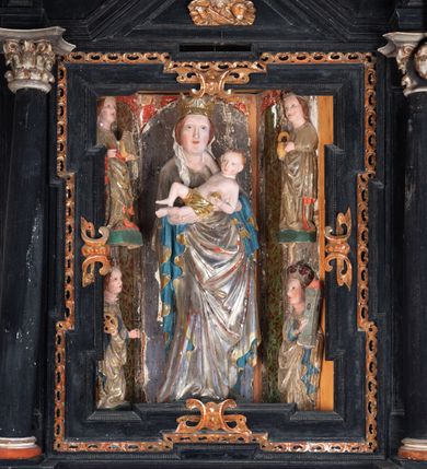 Zdjęcie nr 35: Retabulum ołtarzowe z dużą rzeźbą Matki Boskiej z Dzieciątkiem oraz mniejszymi o połowę czterema rzeźbami Świętych Dziewic umieszczonymi w prostopadłościennej szafie na rzucie trapezu (z ukośnie ustawionymi ściankami bocznymi). Rzeźby pełnoplastyczne, ścięte z tyłu, drążone, ustawione na cokolikach. Pośrodku figura Matki Boskiej w stylu gotyku międzynarodowego; zwrócona en face, w delikatnym kontrapoście, o esowato wygiętej sylwetce, podtrzymuje na obu rękach wpółleżące Dzieciatko. Twarz owalna z podwójnym podbródkiem, rysy twarzy płytko rzeźbione, delikatne, długi, prosty nos. Włosy upięte z tyłu. Maria ubrana w srebrzoną suknię z tkaniną drapowaną po środku w trzy fałdy misowe oraz złocony płaszcz podbity na niebieską podszewką, spływający od tyłu głowy na ramiona i boki, w układzie kaskadowym. Na głowie korona otwarta. Dzieciatko wpółleżące, zwrócone en trois quarts w prawo, prawą ręka ujmuje Matkę za szyję, w lewej trzyma złotą kulę. Twarz okrągła, duże uszy, włosy krótkie, kręcone. W partii bioder przepasany ściśle przylegającą, złotą tkaniną, delikatnie drapowaną. Na lewej ściance szafy u góry figurka św. Cecylii, zwróconej en face, o esowato wygiętej sylwetce, w lewej ręce, opartej na biodrze trzyma piszczałki, prawa wyciągnięta do przodu. Twarz okrągła o pełnych policzkach i płytko rzeźbionych rysach twarzy, włosy długie, falujące fakturowane, spływające przez ramiona na plecy. Ubrana w srebrzoną suknię oraz złocony płaszcz z czerwoną podszewką, spięty na piersi fibulą, z obfitą draperią przewieszoną przez ramiona, opadająca na froncie fałdami misowymi oraz spływającymi kaskadami od ramion. Na głowie blok drewna z namalowaną koroną otwartą.Na lewej ściance u dołu figura św. Katarzyny Aleksandryjskiej, zwróconej en face, w prawej ręce podtrzymuje koło, lewą wyciąga do przodu. Twarz okrągła, policzki pełne, o wysokim czole, nos duży, oczy duże delikatnie zarysowane. Włosy falowane, fakturowane, spływają na plecy. Święta ubrana w srebrzona suknię przepasaną w talii oraz złocony płaszcz spięty broszą na piersi, z obfitą draperią przewieszoną przez ramiona, opadająca na froncie misowymi fałdami i spływającymi kaskadami od ramion. Na głowie blok drewna z namalowaną koroną otwartą. Na prawej ściance u góry figura św. Doroty, zwrócona en face w delikatnym kontrapoście, prawa ręką podtrzymuje koszyk, lewą wskazuje na niego. Twarz owalna, pełna, delikatne rysy, wąski zadarty nos. Włosy falowane, fakturowane, opadają na tył głowy. Ubrana w złotą suknię, delikatnie drapowaną u dołu, oraz srebrzony płaszcz podbity czerwoną podszewką, spięty na piersi i układający się z przodu w fałdy misowe. Na głowie blok drewna z namalowaną koroną otwartą. Na prawej ściance u dołu figura św. Barbary, zwróconej en face, o esowato przegiętej sylwetce, obiema rękami przytrzymuje opartą na lewym ramieniu wieżę. Twarz owalna, pełna, delikatne rysy, wąski zadarty nos. Włosy falowane, fakturowane, opadają na tył głowy. Ubrana w suknię i srebrzony płaszcz z niebieską podszewką, spięty na piersi fibulą, przerzucony przez ramiona z obfitą partią materiału układającą się z przodu w fałdy misowe. Na głowie blok drewna z namalowaną koroną otwartą. Karnacje w tonacji naturalnej, szaty srebrzone i złocone.