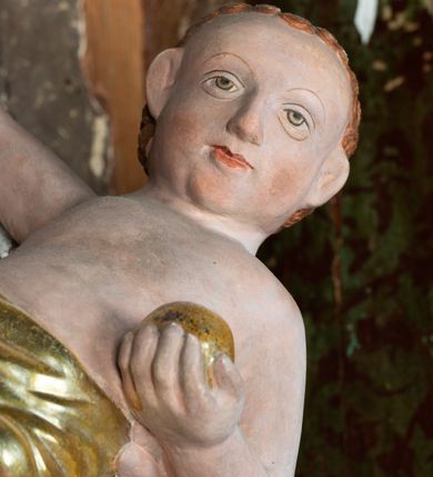 Zdjęcie nr 17: Retabulum ołtarzowe z dużą rzeźbą Matki Boskiej z Dzieciątkiem oraz mniejszymi o połowę czterema rzeźbami Świętych Dziewic umieszczonymi w prostopadłościennej szafie na rzucie trapezu (z ukośnie ustawionymi ściankami bocznymi). Rzeźby pełnoplastyczne, ścięte z tyłu, drążone, ustawione na cokolikach. Pośrodku figura Matki Boskiej w stylu gotyku międzynarodowego; zwrócona en face, w delikatnym kontrapoście, o esowato wygiętej sylwetce, podtrzymuje na obu rękach wpółleżące Dzieciatko. Twarz owalna z podwójnym podbródkiem, rysy twarzy płytko rzeźbione, delikatne, długi, prosty nos. Włosy upięte z tyłu. Maria ubrana w srebrzoną suknię z tkaniną drapowaną po środku w trzy fałdy misowe oraz złocony płaszcz podbity na niebieską podszewką, spływający od tyłu głowy na ramiona i boki, w układzie kaskadowym. Na głowie korona otwarta. Dzieciatko wpółleżące, zwrócone en trois quarts w prawo, prawą ręka ujmuje Matkę za szyję, w lewej trzyma złotą kulę. Twarz okrągła, duże uszy, włosy krótkie, kręcone. W partii bioder przepasany ściśle przylegającą, złotą tkaniną, delikatnie drapowaną. Na lewej ściance szafy u góry figurka św. Cecylii, zwróconej en face, o esowato wygiętej sylwetce, w lewej ręce, opartej na biodrze trzyma piszczałki, prawa wyciągnięta do przodu. Twarz okrągła o pełnych policzkach i płytko rzeźbionych rysach twarzy, włosy długie, falujące fakturowane, spływające przez ramiona na plecy. Ubrana w srebrzoną suknię oraz złocony płaszcz z czerwoną podszewką, spięty na piersi fibulą, z obfitą draperią przewieszoną przez ramiona, opadająca na froncie fałdami misowymi oraz spływającymi kaskadami od ramion. Na głowie blok drewna z namalowaną koroną otwartą.Na lewej ściance u dołu figura św. Katarzyny Aleksandryjskiej, zwróconej en face, w prawej ręce podtrzymuje koło, lewą wyciąga do przodu. Twarz okrągła, policzki pełne, o wysokim czole, nos duży, oczy duże delikatnie zarysowane. Włosy falowane, fakturowane, spływają na plecy. Święta ubrana w srebrzona suknię przepasaną w talii oraz złocony płaszcz spięty broszą na piersi, z obfitą draperią przewieszoną przez ramiona, opadająca na froncie misowymi fałdami i spływającymi kaskadami od ramion. Na głowie blok drewna z namalowaną koroną otwartą. Na prawej ściance u góry figura św. Doroty, zwrócona en face w delikatnym kontrapoście, prawa ręką podtrzymuje koszyk, lewą wskazuje na niego. Twarz owalna, pełna, delikatne rysy, wąski zadarty nos. Włosy falowane, fakturowane, opadają na tył głowy. Ubrana w złotą suknię, delikatnie drapowaną u dołu, oraz srebrzony płaszcz podbity czerwoną podszewką, spięty na piersi i układający się z przodu w fałdy misowe. Na głowie blok drewna z namalowaną koroną otwartą. Na prawej ściance u dołu figura św. Barbary, zwróconej en face, o esowato przegiętej sylwetce, obiema rękami przytrzymuje opartą na lewym ramieniu wieżę. Twarz owalna, pełna, delikatne rysy, wąski zadarty nos. Włosy falowane, fakturowane, opadają na tył głowy. Ubrana w suknię i srebrzony płaszcz z niebieską podszewką, spięty na piersi fibulą, przerzucony przez ramiona z obfitą partią materiału układającą się z przodu w fałdy misowe. Na głowie blok drewna z namalowaną koroną otwartą. Karnacje w tonacji naturalnej, szaty srebrzone i złocone.