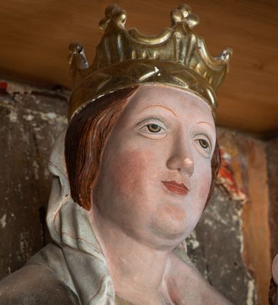 Zdjęcie nr 16: Retabulum ołtarzowe z dużą rzeźbą Matki Boskiej z Dzieciątkiem oraz mniejszymi o połowę czterema rzeźbami Świętych Dziewic umieszczonymi w prostopadłościennej szafie na rzucie trapezu (z ukośnie ustawionymi ściankami bocznymi). Rzeźby pełnoplastyczne, ścięte z tyłu, drążone, ustawione na cokolikach. Pośrodku figura Matki Boskiej w stylu gotyku międzynarodowego; zwrócona en face, w delikatnym kontrapoście, o esowato wygiętej sylwetce, podtrzymuje na obu rękach wpółleżące Dzieciatko. Twarz owalna z podwójnym podbródkiem, rysy twarzy płytko rzeźbione, delikatne, długi, prosty nos. Włosy upięte z tyłu. Maria ubrana w srebrzoną suknię z tkaniną drapowaną po środku w trzy fałdy misowe oraz złocony płaszcz podbity na niebieską podszewką, spływający od tyłu głowy na ramiona i boki, w układzie kaskadowym. Na głowie korona otwarta. Dzieciatko wpółleżące, zwrócone en trois quarts w prawo, prawą ręka ujmuje Matkę za szyję, w lewej trzyma złotą kulę. Twarz okrągła, duże uszy, włosy krótkie, kręcone. W partii bioder przepasany ściśle przylegającą, złotą tkaniną, delikatnie drapowaną. Na lewej ściance szafy u góry figurka św. Cecylii, zwróconej en face, o esowato wygiętej sylwetce, w lewej ręce, opartej na biodrze trzyma piszczałki, prawa wyciągnięta do przodu. Twarz okrągła o pełnych policzkach i płytko rzeźbionych rysach twarzy, włosy długie, falujące fakturowane, spływające przez ramiona na plecy. Ubrana w srebrzoną suknię oraz złocony płaszcz z czerwoną podszewką, spięty na piersi fibulą, z obfitą draperią przewieszoną przez ramiona, opadająca na froncie fałdami misowymi oraz spływającymi kaskadami od ramion. Na głowie blok drewna z namalowaną koroną otwartą.Na lewej ściance u dołu figura św. Katarzyny Aleksandryjskiej, zwróconej en face, w prawej ręce podtrzymuje koło, lewą wyciąga do przodu. Twarz okrągła, policzki pełne, o wysokim czole, nos duży, oczy duże delikatnie zarysowane. Włosy falowane, fakturowane, spływają na plecy. Święta ubrana w srebrzona suknię przepasaną w talii oraz złocony płaszcz spięty broszą na piersi, z obfitą draperią przewieszoną przez ramiona, opadająca na froncie misowymi fałdami i spływającymi kaskadami od ramion. Na głowie blok drewna z namalowaną koroną otwartą. Na prawej ściance u góry figura św. Doroty, zwrócona en face w delikatnym kontrapoście, prawa ręką podtrzymuje koszyk, lewą wskazuje na niego. Twarz owalna, pełna, delikatne rysy, wąski zadarty nos. Włosy falowane, fakturowane, opadają na tył głowy. Ubrana w złotą suknię, delikatnie drapowaną u dołu, oraz srebrzony płaszcz podbity czerwoną podszewką, spięty na piersi i układający się z przodu w fałdy misowe. Na głowie blok drewna z namalowaną koroną otwartą. Na prawej ściance u dołu figura św. Barbary, zwróconej en face, o esowato przegiętej sylwetce, obiema rękami przytrzymuje opartą na lewym ramieniu wieżę. Twarz owalna, pełna, delikatne rysy, wąski zadarty nos. Włosy falowane, fakturowane, opadają na tył głowy. Ubrana w suknię i srebrzony płaszcz z niebieską podszewką, spięty na piersi fibulą, przerzucony przez ramiona z obfitą partią materiału układającą się z przodu w fałdy misowe. Na głowie blok drewna z namalowaną koroną otwartą. Karnacje w tonacji naturalnej, szaty srebrzone i złocone.