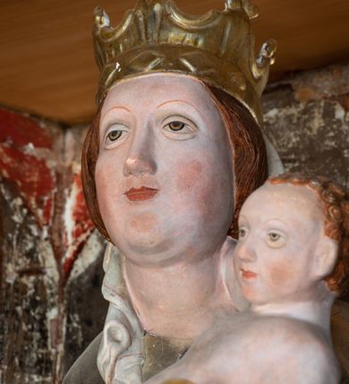 Zdjęcie nr 14: Retabulum ołtarzowe z dużą rzeźbą Matki Boskiej z Dzieciątkiem oraz mniejszymi o połowę czterema rzeźbami Świętych Dziewic umieszczonymi w prostopadłościennej szafie na rzucie trapezu (z ukośnie ustawionymi ściankami bocznymi). Rzeźby pełnoplastyczne, ścięte z tyłu, drążone, ustawione na cokolikach. Pośrodku figura Matki Boskiej w stylu gotyku międzynarodowego; zwrócona en face, w delikatnym kontrapoście, o esowato wygiętej sylwetce, podtrzymuje na obu rękach wpółleżące Dzieciatko. Twarz owalna z podwójnym podbródkiem, rysy twarzy płytko rzeźbione, delikatne, długi, prosty nos. Włosy upięte z tyłu. Maria ubrana w srebrzoną suknię z tkaniną drapowaną po środku w trzy fałdy misowe oraz złocony płaszcz podbity na niebieską podszewką, spływający od tyłu głowy na ramiona i boki, w układzie kaskadowym. Na głowie korona otwarta. Dzieciatko wpółleżące, zwrócone en trois quarts w prawo, prawą ręka ujmuje Matkę za szyję, w lewej trzyma złotą kulę. Twarz okrągła, duże uszy, włosy krótkie, kręcone. W partii bioder przepasany ściśle przylegającą, złotą tkaniną, delikatnie drapowaną. Na lewej ściance szafy u góry figurka św. Cecylii, zwróconej en face, o esowato wygiętej sylwetce, w lewej ręce, opartej na biodrze trzyma piszczałki, prawa wyciągnięta do przodu. Twarz okrągła o pełnych policzkach i płytko rzeźbionych rysach twarzy, włosy długie, falujące fakturowane, spływające przez ramiona na plecy. Ubrana w srebrzoną suknię oraz złocony płaszcz z czerwoną podszewką, spięty na piersi fibulą, z obfitą draperią przewieszoną przez ramiona, opadająca na froncie fałdami misowymi oraz spływającymi kaskadami od ramion. Na głowie blok drewna z namalowaną koroną otwartą.Na lewej ściance u dołu figura św. Katarzyny Aleksandryjskiej, zwróconej en face, w prawej ręce podtrzymuje koło, lewą wyciąga do przodu. Twarz okrągła, policzki pełne, o wysokim czole, nos duży, oczy duże delikatnie zarysowane. Włosy falowane, fakturowane, spływają na plecy. Święta ubrana w srebrzona suknię przepasaną w talii oraz złocony płaszcz spięty broszą na piersi, z obfitą draperią przewieszoną przez ramiona, opadająca na froncie misowymi fałdami i spływającymi kaskadami od ramion. Na głowie blok drewna z namalowaną koroną otwartą. Na prawej ściance u góry figura św. Doroty, zwrócona en face w delikatnym kontrapoście, prawa ręką podtrzymuje koszyk, lewą wskazuje na niego. Twarz owalna, pełna, delikatne rysy, wąski zadarty nos. Włosy falowane, fakturowane, opadają na tył głowy. Ubrana w złotą suknię, delikatnie drapowaną u dołu, oraz srebrzony płaszcz podbity czerwoną podszewką, spięty na piersi i układający się z przodu w fałdy misowe. Na głowie blok drewna z namalowaną koroną otwartą. Na prawej ściance u dołu figura św. Barbary, zwróconej en face, o esowato przegiętej sylwetce, obiema rękami przytrzymuje opartą na lewym ramieniu wieżę. Twarz owalna, pełna, delikatne rysy, wąski zadarty nos. Włosy falowane, fakturowane, opadają na tył głowy. Ubrana w suknię i srebrzony płaszcz z niebieską podszewką, spięty na piersi fibulą, przerzucony przez ramiona z obfitą partią materiału układającą się z przodu w fałdy misowe. Na głowie blok drewna z namalowaną koroną otwartą. Karnacje w tonacji naturalnej, szaty srebrzone i złocone.