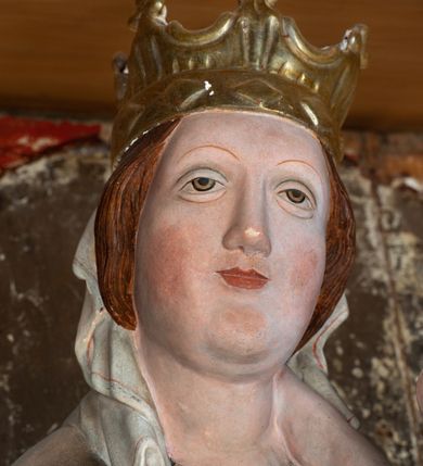 Zdjęcie nr 15: Retabulum ołtarzowe z dużą rzeźbą Matki Boskiej z Dzieciątkiem oraz mniejszymi o połowę czterema rzeźbami Świętych Dziewic umieszczonymi w prostopadłościennej szafie na rzucie trapezu (z ukośnie ustawionymi ściankami bocznymi). Rzeźby pełnoplastyczne, ścięte z tyłu, drążone, ustawione na cokolikach. Pośrodku figura Matki Boskiej w stylu gotyku międzynarodowego; zwrócona en face, w delikatnym kontrapoście, o esowato wygiętej sylwetce, podtrzymuje na obu rękach wpółleżące Dzieciatko. Twarz owalna z podwójnym podbródkiem, rysy twarzy płytko rzeźbione, delikatne, długi, prosty nos. Włosy upięte z tyłu. Maria ubrana w srebrzoną suknię z tkaniną drapowaną po środku w trzy fałdy misowe oraz złocony płaszcz podbity na niebieską podszewką, spływający od tyłu głowy na ramiona i boki, w układzie kaskadowym. Na głowie korona otwarta. Dzieciatko wpółleżące, zwrócone en trois quarts w prawo, prawą ręka ujmuje Matkę za szyję, w lewej trzyma złotą kulę. Twarz okrągła, duże uszy, włosy krótkie, kręcone. W partii bioder przepasany ściśle przylegającą, złotą tkaniną, delikatnie drapowaną. Na lewej ściance szafy u góry figurka św. Cecylii, zwróconej en face, o esowato wygiętej sylwetce, w lewej ręce, opartej na biodrze trzyma piszczałki, prawa wyciągnięta do przodu. Twarz okrągła o pełnych policzkach i płytko rzeźbionych rysach twarzy, włosy długie, falujące fakturowane, spływające przez ramiona na plecy. Ubrana w srebrzoną suknię oraz złocony płaszcz z czerwoną podszewką, spięty na piersi fibulą, z obfitą draperią przewieszoną przez ramiona, opadająca na froncie fałdami misowymi oraz spływającymi kaskadami od ramion. Na głowie blok drewna z namalowaną koroną otwartą.Na lewej ściance u dołu figura św. Katarzyny Aleksandryjskiej, zwróconej en face, w prawej ręce podtrzymuje koło, lewą wyciąga do przodu. Twarz okrągła, policzki pełne, o wysokim czole, nos duży, oczy duże delikatnie zarysowane. Włosy falowane, fakturowane, spływają na plecy. Święta ubrana w srebrzona suknię przepasaną w talii oraz złocony płaszcz spięty broszą na piersi, z obfitą draperią przewieszoną przez ramiona, opadająca na froncie misowymi fałdami i spływającymi kaskadami od ramion. Na głowie blok drewna z namalowaną koroną otwartą. Na prawej ściance u góry figura św. Doroty, zwrócona en face w delikatnym kontrapoście, prawa ręką podtrzymuje koszyk, lewą wskazuje na niego. Twarz owalna, pełna, delikatne rysy, wąski zadarty nos. Włosy falowane, fakturowane, opadają na tył głowy. Ubrana w złotą suknię, delikatnie drapowaną u dołu, oraz srebrzony płaszcz podbity czerwoną podszewką, spięty na piersi i układający się z przodu w fałdy misowe. Na głowie blok drewna z namalowaną koroną otwartą. Na prawej ściance u dołu figura św. Barbary, zwróconej en face, o esowato przegiętej sylwetce, obiema rękami przytrzymuje opartą na lewym ramieniu wieżę. Twarz owalna, pełna, delikatne rysy, wąski zadarty nos. Włosy falowane, fakturowane, opadają na tył głowy. Ubrana w suknię i srebrzony płaszcz z niebieską podszewką, spięty na piersi fibulą, przerzucony przez ramiona z obfitą partią materiału układającą się z przodu w fałdy misowe. Na głowie blok drewna z namalowaną koroną otwartą. Karnacje w tonacji naturalnej, szaty srebrzone i złocone.