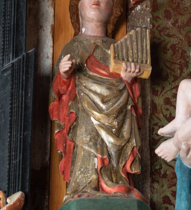 Zdjęcie nr 18: Retabulum ołtarzowe z dużą rzeźbą Matki Boskiej z Dzieciątkiem oraz mniejszymi o połowę czterema rzeźbami Świętych Dziewic umieszczonymi w prostopadłościennej szafie na rzucie trapezu (z ukośnie ustawionymi ściankami bocznymi). Rzeźby pełnoplastyczne, ścięte z tyłu, drążone, ustawione na cokolikach. Pośrodku figura Matki Boskiej w stylu gotyku międzynarodowego; zwrócona en face, w delikatnym kontrapoście, o esowato wygiętej sylwetce, podtrzymuje na obu rękach wpółleżące Dzieciatko. Twarz owalna z podwójnym podbródkiem, rysy twarzy płytko rzeźbione, delikatne, długi, prosty nos. Włosy upięte z tyłu. Maria ubrana w srebrzoną suknię z tkaniną drapowaną po środku w trzy fałdy misowe oraz złocony płaszcz podbity na niebieską podszewką, spływający od tyłu głowy na ramiona i boki, w układzie kaskadowym. Na głowie korona otwarta. Dzieciatko wpółleżące, zwrócone en trois quarts w prawo, prawą ręka ujmuje Matkę za szyję, w lewej trzyma złotą kulę. Twarz okrągła, duże uszy, włosy krótkie, kręcone. W partii bioder przepasany ściśle przylegającą, złotą tkaniną, delikatnie drapowaną. Na lewej ściance szafy u góry figurka św. Cecylii, zwróconej en face, o esowato wygiętej sylwetce, w lewej ręce, opartej na biodrze trzyma piszczałki, prawa wyciągnięta do przodu. Twarz okrągła o pełnych policzkach i płytko rzeźbionych rysach twarzy, włosy długie, falujące fakturowane, spływające przez ramiona na plecy. Ubrana w srebrzoną suknię oraz złocony płaszcz z czerwoną podszewką, spięty na piersi fibulą, z obfitą draperią przewieszoną przez ramiona, opadająca na froncie fałdami misowymi oraz spływającymi kaskadami od ramion. Na głowie blok drewna z namalowaną koroną otwartą.Na lewej ściance u dołu figura św. Katarzyny Aleksandryjskiej, zwróconej en face, w prawej ręce podtrzymuje koło, lewą wyciąga do przodu. Twarz okrągła, policzki pełne, o wysokim czole, nos duży, oczy duże delikatnie zarysowane. Włosy falowane, fakturowane, spływają na plecy. Święta ubrana w srebrzona suknię przepasaną w talii oraz złocony płaszcz spięty broszą na piersi, z obfitą draperią przewieszoną przez ramiona, opadająca na froncie misowymi fałdami i spływającymi kaskadami od ramion. Na głowie blok drewna z namalowaną koroną otwartą. Na prawej ściance u góry figura św. Doroty, zwrócona en face w delikatnym kontrapoście, prawa ręką podtrzymuje koszyk, lewą wskazuje na niego. Twarz owalna, pełna, delikatne rysy, wąski zadarty nos. Włosy falowane, fakturowane, opadają na tył głowy. Ubrana w złotą suknię, delikatnie drapowaną u dołu, oraz srebrzony płaszcz podbity czerwoną podszewką, spięty na piersi i układający się z przodu w fałdy misowe. Na głowie blok drewna z namalowaną koroną otwartą. Na prawej ściance u dołu figura św. Barbary, zwróconej en face, o esowato przegiętej sylwetce, obiema rękami przytrzymuje opartą na lewym ramieniu wieżę. Twarz owalna, pełna, delikatne rysy, wąski zadarty nos. Włosy falowane, fakturowane, opadają na tył głowy. Ubrana w suknię i srebrzony płaszcz z niebieską podszewką, spięty na piersi fibulą, przerzucony przez ramiona z obfitą partią materiału układającą się z przodu w fałdy misowe. Na głowie blok drewna z namalowaną koroną otwartą. Karnacje w tonacji naturalnej, szaty srebrzone i złocone.