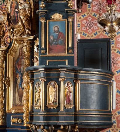 Zdjęcie nr 1: Ambona przyścienna, wisząca, z dostępem przez drzwi od strony zakrystii. Kosz wieloboczny, na cokole, z podwieszeniem w postaci ośmiu kabłąków, zakończonych szyszką. Ścianki kosza wydzielone kolumienkami podtrzymującymi wyłamane na ich osi belkowanie z silnie wysuniętym gzymsem tworzącym parapet. W polach nisze z płaskorzeźbionymi przedstawieniami ewangelistów. Po prawej stronie do kosza dobudowany ćwierćkolisty podest z pełną balustradą. Zaplecek wąski, ujęty dwiema kolumienkami stojącymi na wspornikach, podtrzymującymi imposty, na których stoją wazoniki. W dolnej części płycina. W polu obraz w formie stojącego prostokąta, ujęty ramą zakończoną muszlą, z przedstawieniem Chrystusa w typie Salvator Mundi. Baldachim na planie ośmioboku, w formie belkowania, na którym ustawione są kabłąki, podtrzymujące figurę anioła grającego na trąbie. Na podniebiu baldachimu figura gołębicy Ducha Świętego w glorii promienistej. Struktura polichromowana w kolorze niebiesko-zielonym; kolumienki, profile i ornamentyka złocone. 
