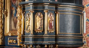 Zdjęcie nr 1: Ambona przyścienna, wisząca, z dostępem przez drzwi od strony zakrystii. Kosz wieloboczny, na cokole, z podwieszeniem w postaci ośmiu kabłąków, zakończonych szyszką. Ścianki kosza wydzielone kolumienkami podtrzymującymi wyłamane na ich osi belkowanie z silnie wysuniętym gzymsem tworzącym parapet. W polach nisze z płaskorzeźbionymi przedstawieniami ewangelistów. Po prawej stronie do kosza dobudowany ćwierćkolisty podest z pełną balustradą. Zaplecek wąski, ujęty dwiema kolumienkami stojącymi na wspornikach, podtrzymującymi imposty, na których stoją wazoniki. W dolnej części płycina. W polu obraz w formie stojącego prostokąta, ujęty ramą zakończoną muszlą, z przedstawieniem Chrystusa w typie Salvator Mundi. Baldachim na planie ośmioboku, w formie belkowania, na którym ustawione są kabłąki, podtrzymujące figurę anioła grającego na trąbie. Na podniebiu baldachimu figura gołębicy Ducha Świętego w glorii promienistej. Struktura polichromowana w kolorze niebiesko-zielonym; kolumienki, profile i ornamentyka złocone. 
