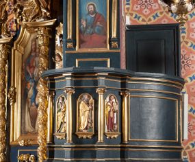 Zdjęcie nr 1: Ambona przyścienna, wisząca, z dostępem przez drzwi od strony zakrystii. Kosz wieloboczny, na cokole, z podwieszeniem w postaci ośmiu kabłąków, zakończonych szyszką. Ścianki kosza wydzielone kolumienkami podtrzymującymi wyłamane na ich osi belkowanie z silnie wysuniętym gzymsem tworzącym parapet. W polach nisze z płaskorzeźbionymi przedstawieniami ewangelistów. Po prawej stronie do kosza dobudowany ćwierćkolisty podest z pełną balustradą. Zaplecek wąski, ujęty dwiema kolumienkami stojącymi na wspornikach, podtrzymującymi imposty, na których stoją wazoniki. W dolnej części płycina. W polu obraz w formie stojącego prostokąta, ujęty ramą zakończoną muszlą, z przedstawieniem Chrystusa w typie Salvator Mundi. Baldachim na planie ośmioboku, w formie belkowania, na którym ustawione są kabłąki, podtrzymujące figurę anioła grającego na trąbie. Na podniebiu baldachimu figura gołębicy Ducha Świętego w glorii promienistej. Struktura polichromowana w kolorze niebiesko-zielonym; kolumienki, profile i ornamentyka złocone. 
