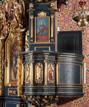 Zdjęcie nr 1: Ambona przyścienna, wisząca, z dostępem przez drzwi od strony zakrystii. Kosz wieloboczny, na cokole, z podwieszeniem w postaci ośmiu kabłąków, zakończonych szyszką. Ścianki kosza wydzielone kolumienkami podtrzymującymi wyłamane na ich osi belkowanie z silnie wysuniętym gzymsem tworzącym parapet. W polach nisze z płaskorzeźbionymi przedstawieniami ewangelistów. Po prawej stronie do kosza dobudowany ćwierćkolisty podest z pełną balustradą. Zaplecek wąski, ujęty dwiema kolumienkami stojącymi na wspornikach, podtrzymującymi imposty, na których stoją wazoniki. W dolnej części płycina. W polu obraz w formie stojącego prostokąta, ujęty ramą zakończoną muszlą, z przedstawieniem Chrystusa w typie Salvator Mundi. Baldachim na planie ośmioboku, w formie belkowania, na którym ustawione są kabłąki, podtrzymujące figurę anioła grającego na trąbie. Na podniebiu baldachimu figura gołębicy Ducha Świętego w glorii promienistej. Struktura polichromowana w kolorze niebiesko-zielonym; kolumienki, profile i ornamentyka złocone. 
