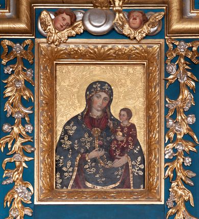 Zdjęcie nr 2: Obraz w formie stojącego prostokąta. W centrum Matka Boska ukazana w półpostaci, frontalnie, z głową lekko przechyloną w lewo. W prawej dłoni, na tle piersi trzyma kwiat róży, lewą podtrzymuje na ramieniu Dzieciątko. Twarz ma pociągłą, o migdałowatych oczach, prostym nosie i małych, pełnych ustach, okoloną jasnobrązowymi, kręconymi włosami ukrytymi pod welonem. Ubrana jest w czerwoną suknię o dekoracyjnej, złoconej lamówce z malowaną imitacją kameryzacji. Na głowę i ramiona ma narzucony ciemnoniebieski maforion o złoconym wzorze kwiatowym, z dekoracyjną, złoconą lamówką, z malowaną imitacją kameryzacji, na piersi zapięty okrągłą broszą z malowanym kamieniem szlachetnym. Dzieciątko w pozycji siedzącej, zwrócone w trzech czwartych w prawo, lewą ręką podtrzymuje złocone jabłko królewskie, prawą unosi w geście błogosławieństwa. Twarz ma szczupłą, trójkątną, o dużych, migdałowatych oczach, małym nosie i ustach, okoloną jasnobrązowymi, krótkimi włosami. Ubrane jest w czerwoną suknię o złoconym wzorze kwiatowym, ze złoconą lamówką z imitacją kameryzacji. Postacie ukazane na złoconym tle, z rytym w zaprawie wzorem wici roślinnej oraz rytymi w zaprawie nimbami wokół ich głów. 
