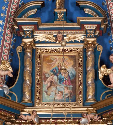 Zdjęcie nr 1: Obraz w formie stojącego prostokąta. W centrum na obłokach ukazana siedząca Maria, zwrócona w trzech czwartych w lewo, prawą rękę opiera na obłoku, lewą obejmuje Dzieciątko siedzące na jej kolanach. Twarz ma okrągłą, o migdałowatych oczach, prostym nosie i małych ustach, okoloną brązowymi włosami, opadającymi na ramiona. Ubrana jest w ciemnoróżową suknię oraz niebieski płaszcz zarzucony na ramiona i nogi, na głowie ma żółty welon. Dzieciątko w pozycji siedzącej, zwrócone w trzech czwartych w lewo, unosi wysoko prawą rękę w geście błogosławieństwa. Twarz ma okrągłą, o pełnych policzkach i delikatnych rysach, okoloną krótkimi, brązowymi włosami. Ukazane jest jako nagie, o pulchnym ciele. Nad głową Marii dwa putta podtrzymują otwartą koronę. W dolnej części obrazu trzy putta pośród obłoków trzymające kwiaty i wieńce, z których sypią płatki. Postacie ukazane na tle pierzastych chmur, żółto-brązowych w górnej części i niebiesko-szarych w dolnej części obrazu. W lewym dolnym rogu obrazu sygnatura „A(leksandra) Adamska-Kuś”.
