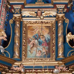 Zdjęcie nr 1: Obraz w formie stojącego prostokąta. W centrum na obłokach ukazana siedząca Maria, zwrócona w trzech czwartych w lewo, prawą rękę opiera na obłoku, lewą obejmuje Dzieciątko siedzące na jej kolanach. Twarz ma okrągłą, o migdałowatych oczach, prostym nosie i małych ustach, okoloną brązowymi włosami, opadającymi na ramiona. Ubrana jest w ciemnoróżową suknię oraz niebieski płaszcz zarzucony na ramiona i nogi, na głowie ma żółty welon. Dzieciątko w pozycji siedzącej, zwrócone w trzech czwartych w lewo, unosi wysoko prawą rękę w geście błogosławieństwa. Twarz ma okrągłą, o pełnych policzkach i delikatnych rysach, okoloną krótkimi, brązowymi włosami. Ukazane jest jako nagie, o pulchnym ciele. Nad głową Marii dwa putta podtrzymują otwartą koronę. W dolnej części obrazu trzy putta pośród obłoków trzymające kwiaty i wieńce, z których sypią płatki. Postacie ukazane na tle pierzastych chmur, żółto-brązowych w górnej części i niebiesko-szarych w dolnej części obrazu. W lewym dolnym rogu obrazu sygnatura „A(leksandra) Adamska-Kuś”.
