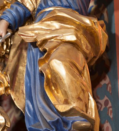 Zdjęcie nr 5: Pełnoplastyczna rzeźba ustawiona na nieregularnej podstawie. Święty ukazany frontalnie, w kontrapoście, z ugiętą lewą nogą oraz pochyloną głową. Lewą rękę unosi wysoko, z wskazującym gestem dłoni, prawą wyciąga w dół, w dłoni trzyma srebrzony miecz, skierowany ostrzem w dół. Twarz ma pociągłą, szczupłą, o migdałowatych oczach, dużym nosie i rozchylonych, pełnych ustach. Okolona jest gęstą, skręconą brodą opadającą na pierś oraz krótkimi, falistymi włosami. Święty ubrany jest w niebieską tunikę, przylegającą do ciała i silnie drapowaną, przepasaną w talii oraz złocony płaszcz przewiązany przez prawe ramię i zawinięty na lewej nodze, na stopach ma złocone buty. Polichromia ciała naturalistyczna.
