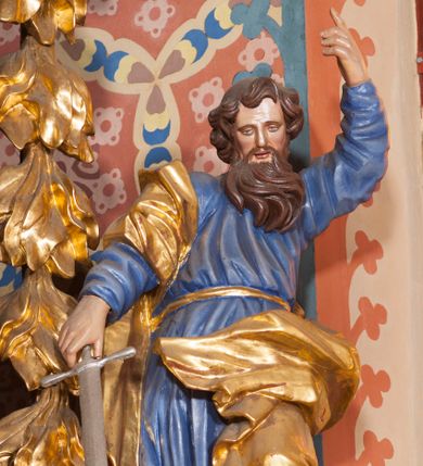 Zdjęcie nr 1: Pełnoplastyczna rzeźba ustawiona na nieregularnej podstawie. Święty ukazany frontalnie, w kontrapoście, z ugiętą lewą nogą oraz pochyloną głową. Lewą rękę unosi wysoko, z wskazującym gestem dłoni, prawą wyciąga w dół, w dłoni trzyma srebrzony miecz, skierowany ostrzem w dół. Twarz ma pociągłą, szczupłą, o migdałowatych oczach, dużym nosie i rozchylonych, pełnych ustach. Okolona jest gęstą, skręconą brodą opadającą na pierś oraz krótkimi, falistymi włosami. Święty ubrany jest w niebieską tunikę, przylegającą do ciała i silnie drapowaną, przepasaną w talii oraz złocony płaszcz przewiązany przez prawe ramię i zawinięty na lewej nodze, na stopach ma złocone buty. Polichromia ciała naturalistyczna.
