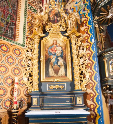 Zdjęcie nr 2: Nastawa ołtarzowa architektoniczna, jednoosiowa, jednokondygnacyjna ze zwieńczeniem. Ołtarz sarkofagowy, wybrzuszony w górnej części, zwężający się ku dołowi, na cokole; mensa drewniana. Na frontowej ścianie skrzyni ołtarzowej płycina w formie odwróconego trapezu. Nastawa ustawiona na dwustrefowym cokole, z wyłamanymi dwoma prostopadłościanami, ujęta parą kolumn o kręconych trzonach oplecionych wicią roślinną, dźwigających fragmenty gzymsu wyłamane pod kątem o wklęsłych bokach. Na nich ustawione figury dwóch aniołów. Niemal całe pole główne wypełnione jest wnęką w formie stojącego prostokąta o wklęsłych górnych narożach, z uskokiem, ujętą grubą, profilowaną ramą dekorowaną kwiatami, z obrazem Matki Boskiej Różańcowej. Pod wnęką płycina wypełniona kwiatami. Kondygnacja flankowana uszami utworzonymi z suchego akantu z dwiema parami puttów. Zwieńczenie w formie medalionu z obrazem Zwiastowania ujętego suchym akantem, zakończone cokołem z figurą anioła. Struktura polichromowana w kolorze niebieskim; kolumny, profile i ornamentyka złocone. 