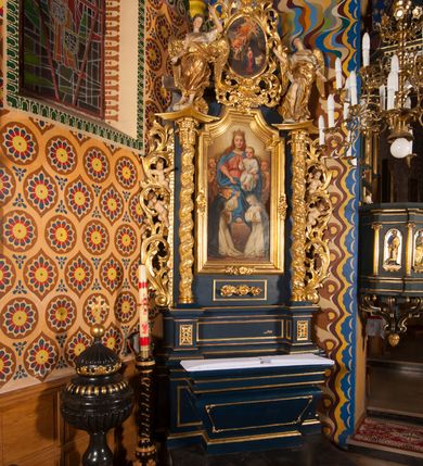 Zdjęcie nr 8: Nastawa ołtarzowa architektoniczna, jednoosiowa, jednokondygnacyjna ze zwieńczeniem. Ołtarz sarkofagowy, wybrzuszony w górnej części, zwężający się ku dołowi, na cokole; mensa drewniana. Na frontowej ścianie skrzyni ołtarzowej płycina w formie odwróconego trapezu. Nastawa ustawiona na dwustrefowym cokole, z wyłamanymi dwoma prostopadłościanami, ujęta parą kolumn o kręconych trzonach oplecionych wicią roślinną, dźwigających fragmenty gzymsu wyłamane pod kątem o wklęsłych bokach. Na nich ustawione figury dwóch aniołów. Niemal całe pole główne wypełnione jest wnęką w formie stojącego prostokąta o wklęsłych górnych narożach, z uskokiem, ujętą grubą, profilowaną ramą dekorowaną kwiatami, z obrazem Matki Boskiej Różańcowej. Pod wnęką płycina wypełniona kwiatami. Kondygnacja flankowana uszami utworzonymi z suchego akantu z dwiema parami puttów. Zwieńczenie w formie medalionu z obrazem Zwiastowania ujętego suchym akantem, zakończone cokołem z figurą anioła. Struktura polichromowana w kolorze niebieskim; kolumny, profile i ornamentyka złocone. 