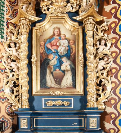 Zdjęcie nr 1: Nastawa ołtarzowa architektoniczna, jednoosiowa, jednokondygnacyjna ze zwieńczeniem. Ołtarz sarkofagowy, wybrzuszony w górnej części, zwężający się ku dołowi, na cokole; mensa drewniana. Na frontowej ścianie skrzyni ołtarzowej płycina w formie odwróconego trapezu. Nastawa ustawiona na dwustrefowym cokole, z wyłamanymi dwoma prostopadłościanami, ujęta parą kolumn o kręconych trzonach oplecionych wicią roślinną, dźwigających fragmenty gzymsu wyłamane pod kątem o wklęsłych bokach. Na nich ustawione figury dwóch aniołów. Niemal całe pole główne wypełnione jest wnęką w formie stojącego prostokąta o wklęsłych górnych narożach, z uskokiem, ujętą grubą, profilowaną ramą dekorowaną kwiatami, z obrazem Matki Boskiej Różańcowej. Pod wnęką płycina wypełniona kwiatami. Kondygnacja flankowana uszami utworzonymi z suchego akantu z dwiema parami puttów. Zwieńczenie w formie medalionu z obrazem Zwiastowania ujętego suchym akantem, zakończone cokołem z figurą anioła. Struktura polichromowana w kolorze niebieskim; kolumny, profile i ornamentyka złocone. 