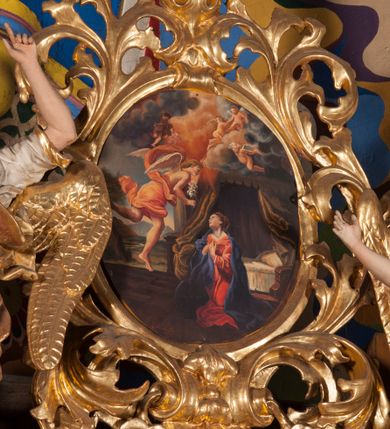 Zdjęcie nr 1: Obraz w formie stojącego owalu. W prawej części ukazana klęcząca Maria, zwrócona w trzech czwartych w prawo, z głową skierowana w górę i dłońmi skrzyżowanymi na piersi. Twarz ma młodzieńczą, o delikatnych rysach, okoloną jasnobrązowymi włosami upiętymi z tyłu głowy. Ubrana jest w czerwoną suknię oraz niebieski płaszcz zarzucony na ramiona. Przed nią, w lewej części obrazu nadlatujący archanioł ukazany z profilu. W prawej ręce trzyma gałązkę lilii, lewą unosi, z wskazującym gestem dłoni. Twarz ma młodzieńczą, okoloną jasnymi włosami. W partii bioder przewiązany jest rozwianą, pomarańczową szatą. Skrzydła ma białe, na wpół rozłożone. Scena ukazana jest we wnętrzu z widocznym po prawej stronie łóżkiem z baldachimem i stojącą obok kolumną, przesłoniętymi w górnej części gęstymi obłokami z puttami. W lewej części obrazu, w oddali widoczny górzysty pejzaż. W dolnej zaś sygnatura „A(leksandra) Adamska-Kuś / [...]”.
