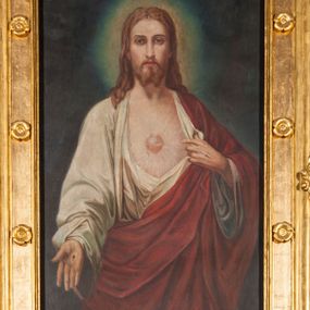 Zdjęcie nr 1: Obraz w formie stojącego prostokąta. W centrum Chrystus ukazany do kolan, frontalnie, lewą ręką odsłania szatę z piersi, prawą wyciąga do przodu, w geście otwartej dłoni. Twarz ma pociągłą, o dużych oczach, długim nosie i pełnych ustach, okoloną krótką, rozdwajającą się brodą oraz jasnymi włosami opadającymi na ramiona. Wokół głowy nimb. Ubrany jest w białą, luźną tunikę, odsłaniającą tors oraz czerwony płaszcz przewiązany diagonalnie przez lewe ramię i otaczający postać. Na jego piersi unosi się gorejące serce w glorii promienistej. Na dłoniach ma ślady męki. Chrystus ukazany jest na cieniowanym zielono-granatowym tle. Obraz ujęty złoconą, szeroką ramą, dekorowaną rzeźbionymi rozetami. 
