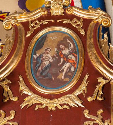 Zdjęcie nr 1: Obraz w formie stojącego owalu z przedstawieniem archanioła Gabriela zwiastującego Marii. Po lewej stronie kompozycji ukazana klęcząca Matka Boska, zwrócona w trzech czwartych w prawo, z rekami złożonymi na piersi oraz głową skierowaną w lewo. Twarz ma owalną o schematycznie zaznaczonych rysach, okoloną brązowymi włosami. Ubrana jest w jasnoróżową suknię, przepasaną w talii oraz niebieski płaszcz zarzucony na prawe ramę i lewy bok, na głowie ma biały welon. Przed Marią leży na klęczniku otwarta księga. W prawej części obrazu archanioł ukazany frontalnie, przechylony w prawo, klęczący na lewe kolano na obłoku, z prawą nogą wyciągniętą w przód. W prawej, opuszczonej wzdłuż ciała ręce trzyma gałązkę lilii, lewą unosi w górę, z dłonią w geście wskazującym. Twarz ma owalną, młodzieńczą, o schematycznie zaznaczonych rysach, okoloną krótkimi, brązowymi włosami. Ubrany jest w białą tunikę o wzorze kwiatowym, przepasaną w talii, odsłaniającą prawą nogę oraz czerwony płaszcz przerzucony przez prawe ramię. Skrzydła ma białe, złożone. Nad postaciami unosi się biała gołębica Ducha Świętego. Scena ukazana jest na tle pejzażu z żółto-szarym niebem. W lewej części obrazu podwieszona zielona kurtyna.

