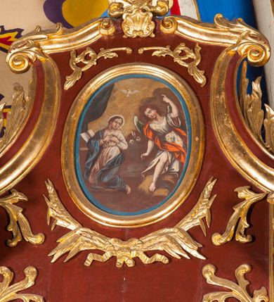 Zdjęcie nr 1: Obraz w formie stojącego owalu z przedstawieniem archanioła Gabriela zwiastującego Marii. Po lewej stronie kompozycji ukazana klęcząca Matka Boska, zwrócona w trzech czwartych w prawo, z rekami złożonymi na piersi oraz głową skierowaną w lewo. Twarz ma owalną o schematycznie zaznaczonych rysach, okoloną brązowymi włosami. Ubrana jest w jasnoróżową suknię, przepasaną w talii oraz niebieski płaszcz zarzucony na prawe ramę i lewy bok, na głowie ma biały welon. Przed Marią leży na klęczniku otwarta księga. W prawej części obrazu archanioł ukazany frontalnie, przechylony w prawo, klęczący na lewe kolano na obłoku, z prawą nogą wyciągniętą w przód. W prawej, opuszczonej wzdłuż ciała ręce trzyma gałązkę lilii, lewą unosi w górę, z dłonią w geście wskazującym. Twarz ma owalną, młodzieńczą, o schematycznie zaznaczonych rysach, okoloną krótkimi, brązowymi włosami. Ubrany jest w białą tunikę o wzorze kwiatowym, przepasaną w talii, odsłaniającą prawą nogę oraz czerwony płaszcz przerzucony przez prawe ramię. Skrzydła ma białe, złożone. Nad postaciami unosi się biała gołębica Ducha Świętego. Scena ukazana jest na tle pejzażu z żółto-szarym niebem. W lewej części obrazu podwieszona zielona kurtyna.


