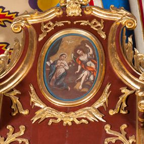 Zdjęcie nr 1: Obraz w formie stojącego owalu z przedstawieniem archanioła Gabriela zwiastującego Marii. Po lewej stronie kompozycji ukazana klęcząca Matka Boska, zwrócona w trzech czwartych w prawo, z rekami złożonymi na piersi oraz głową skierowaną w lewo. Twarz ma owalną o schematycznie zaznaczonych rysach, okoloną brązowymi włosami. Ubrana jest w jasnoróżową suknię, przepasaną w talii oraz niebieski płaszcz zarzucony na prawe ramę i lewy bok, na głowie ma biały welon. Przed Marią leży na klęczniku otwarta księga. W prawej części obrazu archanioł ukazany frontalnie, przechylony w prawo, klęczący na lewe kolano na obłoku, z prawą nogą wyciągniętą w przód. W prawej, opuszczonej wzdłuż ciała ręce trzyma gałązkę lilii, lewą unosi w górę, z dłonią w geście wskazującym. Twarz ma owalną, młodzieńczą, o schematycznie zaznaczonych rysach, okoloną krótkimi, brązowymi włosami. Ubrany jest w białą tunikę o wzorze kwiatowym, przepasaną w talii, odsłaniającą prawą nogę oraz czerwony płaszcz przerzucony przez prawe ramię. Skrzydła ma białe, złożone. Nad postaciami unosi się biała gołębica Ducha Świętego. Scena ukazana jest na tle pejzażu z żółto-szarym niebem. W lewej części obrazu podwieszona zielona kurtyna.

