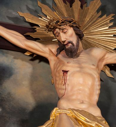 Zdjęcie nr 3: Figura Chrystusa umarłego przybita jest trzema gwoździami do krzyża o prostych ramionach. Ciało w lekkim zwisie o horyzontalnie rozłożonych ramionach, z głową opadającą na prawy bark, nogi silnie ugięte w kolanach, stopy skrzyżowane w układzie prawa na lewą. Chrystus ma podłużną twarz, o migdałowatych, zamkniętych oczach, dużym, garbatym nosie oraz szerokich, rozchylonych ustach, okoloną krótką brodą oraz skręconymi w pukle włosami opadającymi na prawe ramię. Na głowie ma nałożoną koronę cierniową. Ciało ma silnie wychudzone z wyraźnie podkreślonymi guzowatymi żebrami, mostkiem i kośćmi biodrowymi, z silnie wciętą talią oraz zapadłą płaszczyzną brzucha. Nogi i ręce o podkreślonej wklęsło-wypukłej linii mięśni i ścięgien. Złocone perizonium zawieszone na podwójnym sznurze, odsłaniające prawe udo, z rozwianym zwisem tkaniny na prawym boku. Na krzyżowaniu belek krzyża, na tle głowy Zbawiciela gloria promienista. Na zakończeniu pionowej belki krzyża banderola z napisem „IN RI”. Polichromia ciała Chrystusa naturalistyczna, z zaznaczonymi śladami męki. 


