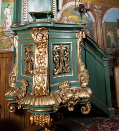 Zdjęcie nr 1: Ambona przyścienna, wisząca. Kosz na planie kwadratu o wklęsłych bokach i ściętych narożach, na profilowanej podstawie dekorowanej muszlą i kartuszem. U jego spodu podwieszone cztery spływy wolutowe z chrząstką i liściem akantu, połączone pośrodku ażurową szyszką. Kosz ujęty w narożach spływami wolutowymi dekorowanymi ornamentem rocaille'owym i uskrzydlonymi główkami anielskimi. Ściany zdobione płycinami z motywem suchego akantu. Parapet w formie gzymsu o wklęsłych bokach i zaokrąglonych narożach z pulpitem. Od wschodu jednobiegowe schody z opilastrowaną balustradą zakończoną niepełnym belkowaniem. Zaplecek wysoki, ujęty dwoma pilastrami, z trzonami ozdobionymi kampanulami, o kapitelach w formie konsol podtrzymujących pełne belkowanie wyłamane na ich osi, ze zdwojonym, przerwanym i odwróconym na zewnątrz przyczółkiem. W polu zaplecka płaskorzeźba z przedstawieniem Chrystusa Dobrego Pasterza. Całość zwieńczona glorią promienistą z gołębicą Ducha Świętego. Struktura polichromowana w kolorze jasnozielonym, profile oraz ornamentyka pozłacane i posrebrzane. 