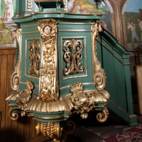 Zdjęcie nr 1: Ambona przyścienna, wisząca. Kosz na planie kwadratu o wklęsłych bokach i ściętych narożach, na profilowanej podstawie dekorowanej muszlą i kartuszem. U jego spodu podwieszone cztery spływy wolutowe z chrząstką i liściem akantu, połączone pośrodku ażurową szyszką. Kosz ujęty w narożach spływami wolutowymi dekorowanymi ornamentem rocaille'owym i uskrzydlonymi główkami anielskimi. Ściany zdobione płycinami z motywem suchego akantu. Parapet w formie gzymsu o wklęsłych bokach i zaokrąglonych narożach z pulpitem. Od wschodu jednobiegowe schody z opilastrowaną balustradą zakończoną niepełnym belkowaniem. Zaplecek wysoki, ujęty dwoma pilastrami, z trzonami ozdobionymi kampanulami, o kapitelach w formie konsol podtrzymujących pełne belkowanie wyłamane na ich osi, ze zdwojonym, przerwanym i odwróconym na zewnątrz przyczółkiem. W polu zaplecka płaskorzeźba z przedstawieniem Chrystusa Dobrego Pasterza. Całość zwieńczona glorią promienistą z gołębicą Ducha Świętego. Struktura polichromowana w kolorze jasnozielonym, profile oraz ornamentyka pozłacane i posrebrzane. 