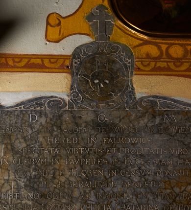 Zdjęcie nr 2: Epitafium w formie leżącego prostokąta, ujęte ramą utworzoną ze zwróconych ku sobie par wolut. U spodu tablicy herb z labrami, zwieńczony klejnotem w postaci hełmu w koronie i zbrojnej nogi, o czteropolowej tarczy z herbami: Nowina, Topór, Kietlicz, Doliwa. Epitafium zwieńczone rytowanym medalionem z przedstawieniem czaszki, skrzyżowanych piszczeli i klepsydry, ujętym ornamentem roślinnym przechodzącym w woluty, zakończonym krzyżem. W polu inskrypcja „D(EO) O(PTIMO) M(AXIMO) / G(E)N(ER)OSO OLIM IACOBO DE WILKOWICE WIEYSKI / HEREDI IN FAŁKOWICE / SPECTATAE VIRTVTIS ET PROBITATIS VIRO / IN CLERVM IN PAVPERES IN ECCLESIAM HANC / CVI MILLE FLOREN(ORUM) IN CENSVM DONAVIT / PIA LIBERALITATE BENEFICO / CHRISTIANO OBITV AD IMMORTALEM VITAM TRANSLATO / MAESTISSIMA CONIVNX RELICTA CHATARINA DE POREMBA / MONVMENTVM HOC POSVIT/ OBIIT ANNO D(OMI)NI 1673 MENSE FEBRVARY DIE 22 / AETATIS SVAE 49.”