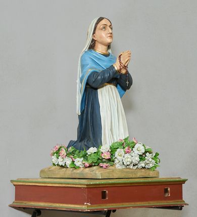 Zdjęcie nr 1: Rzeźba pełna, na podstawie imitującej skałę. Zamontowana na wysokiej podstawie na planie zbliżonym do kwadratu, o krawędziach ujętych profilowanymi listewkami, z dwoma otworami na drążki. Bernadeta ukazana w pozycji klęczącej, z rękami złożonymi w geście modlitwy, z lekko uniesioną głową. Twarz ma pociągłą, o migdałowatych oczach skierowanych w górę, dużym nosie i małych ustach; okoloną długimi brązowymi włosami opadającymi na plecy. Ubrana jest w granatową suknię, o złoconych lamówkach, przepasaną w talii białym fartuchem, na ramiona i plecy ma narzuconą niebieską chustę o złoconych lamówkach, na głowie ma biały welon. Polichromia ciała naturalistyczna. Wokół nóg figury zamocowane sztuczne kwiaty.



